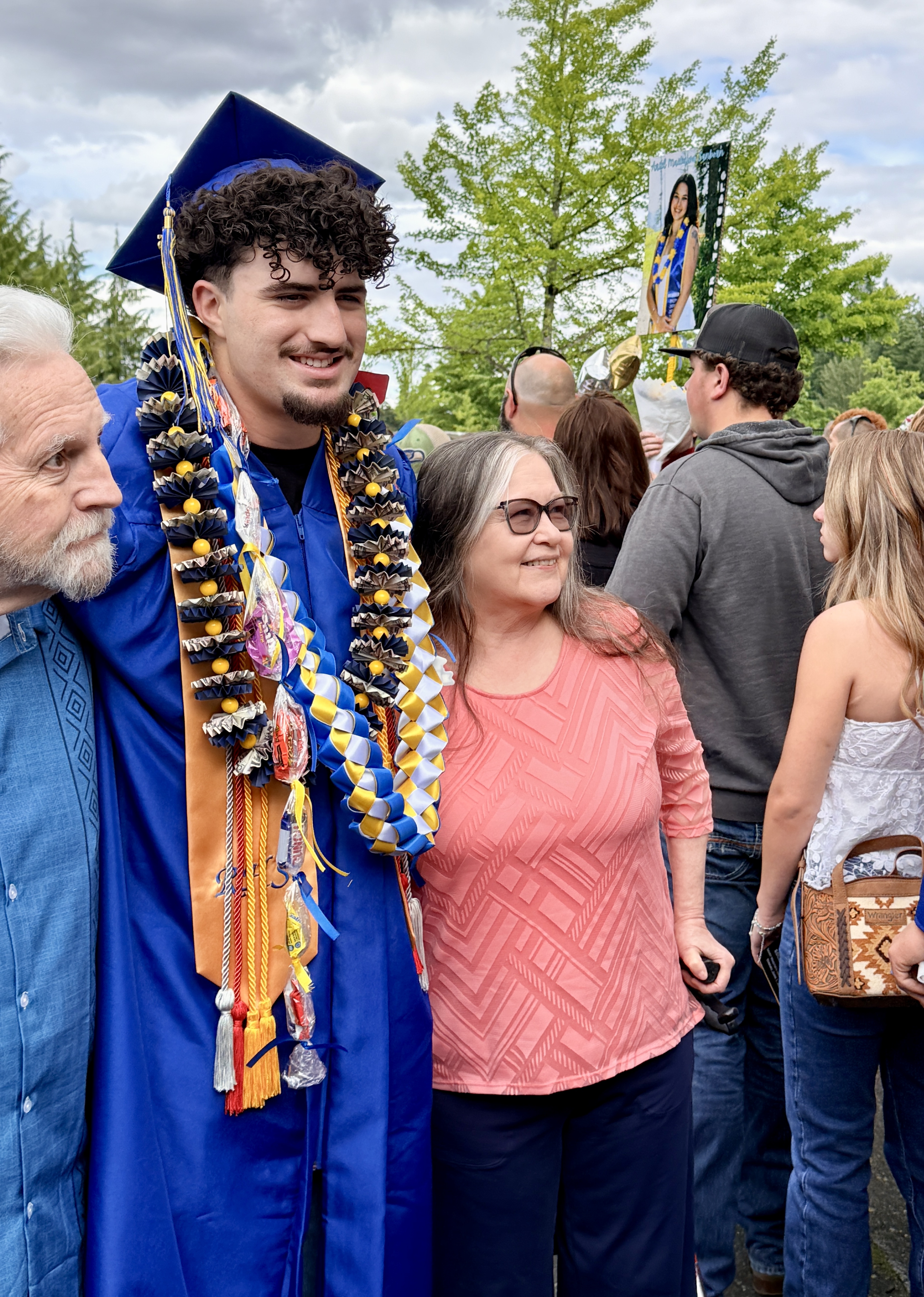A man and woman in a crowd. He wears a graduation gown, cap, and lei. She wears glasses and a patterned top.