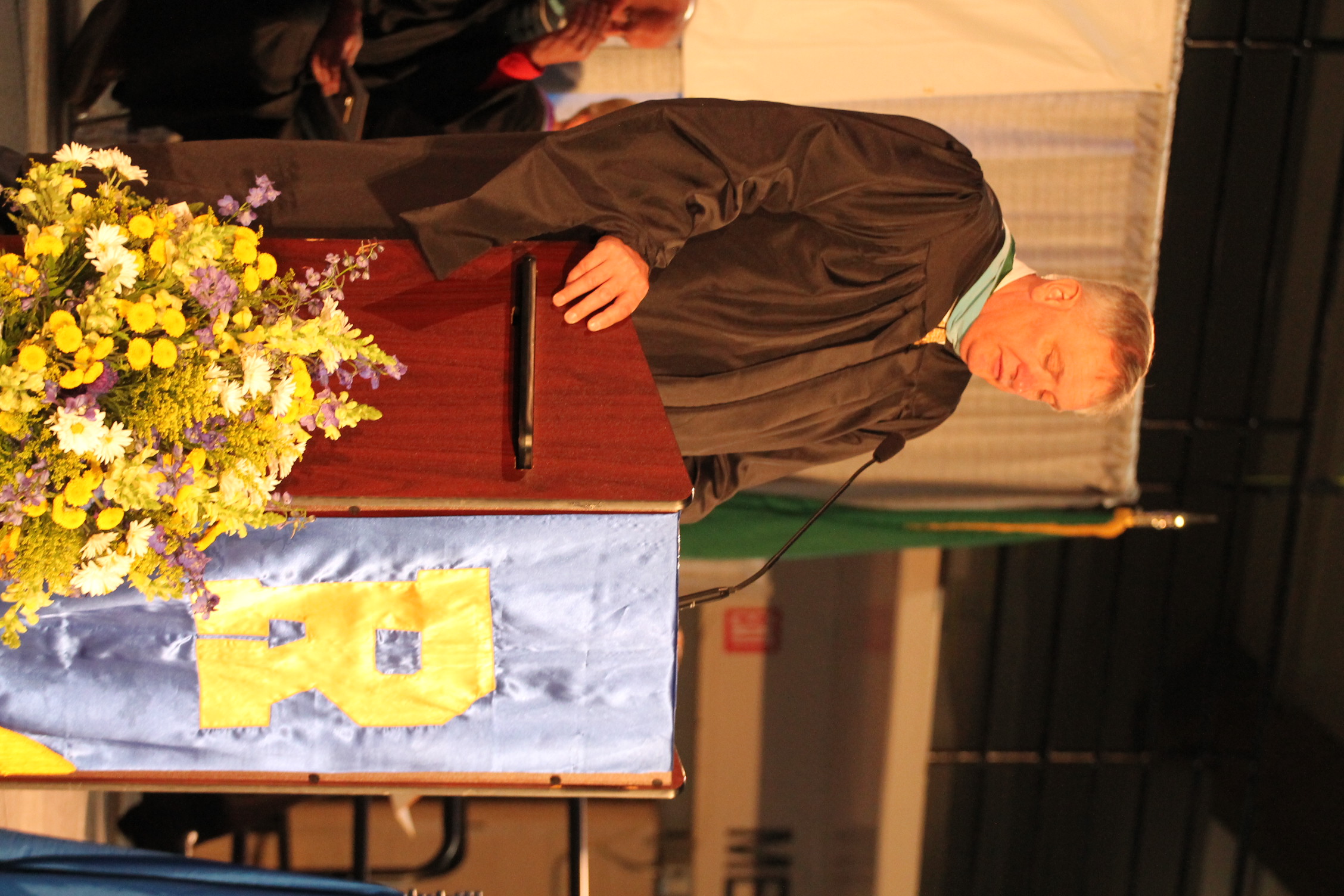 A man in a cap and gown stands at a podium with a microphone and flowers. Behind him, a banner displays a letter "R".