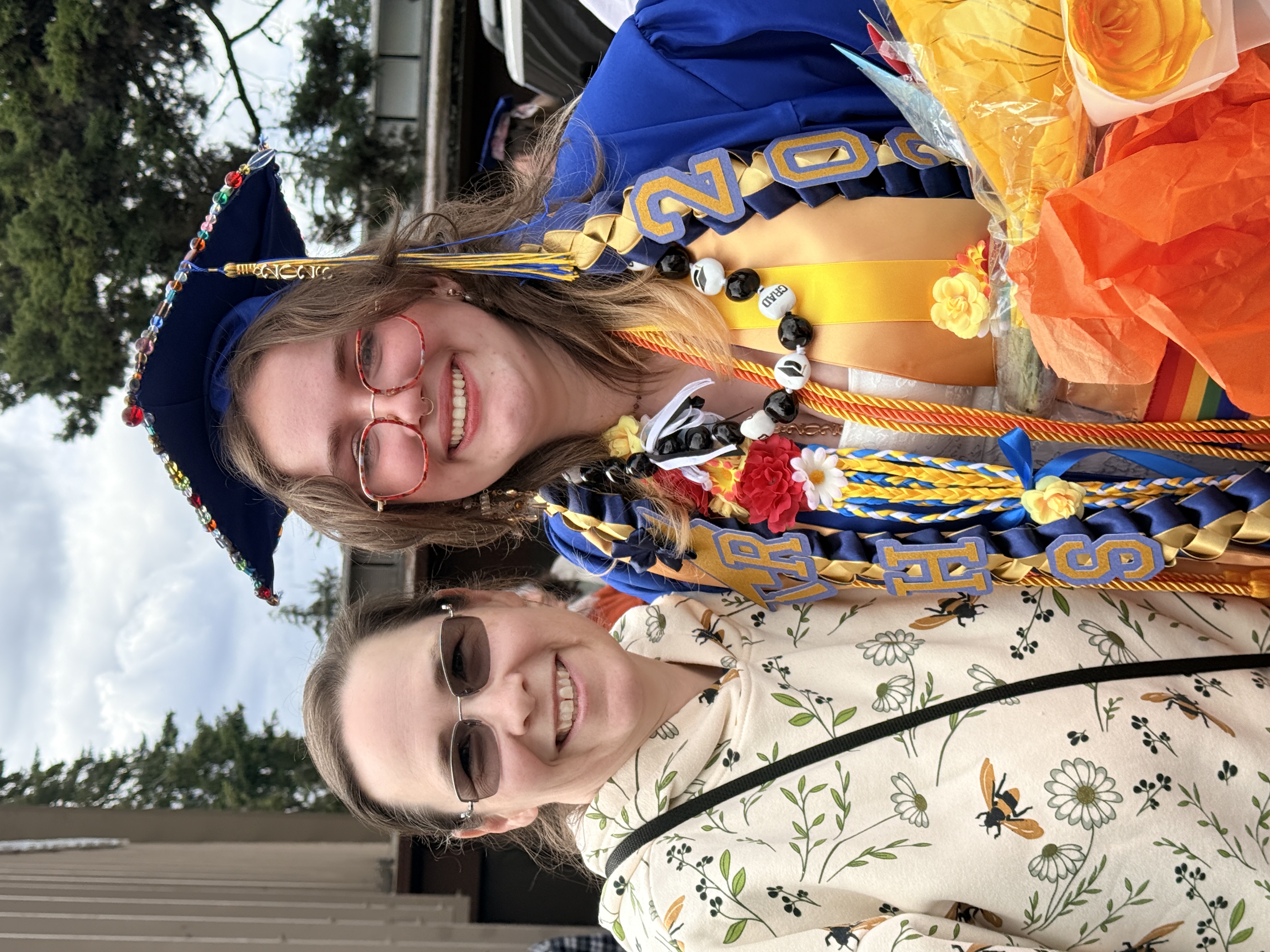Two women stand outside, one in a cap and gown with a bouquet, the other in a floral hoodie.