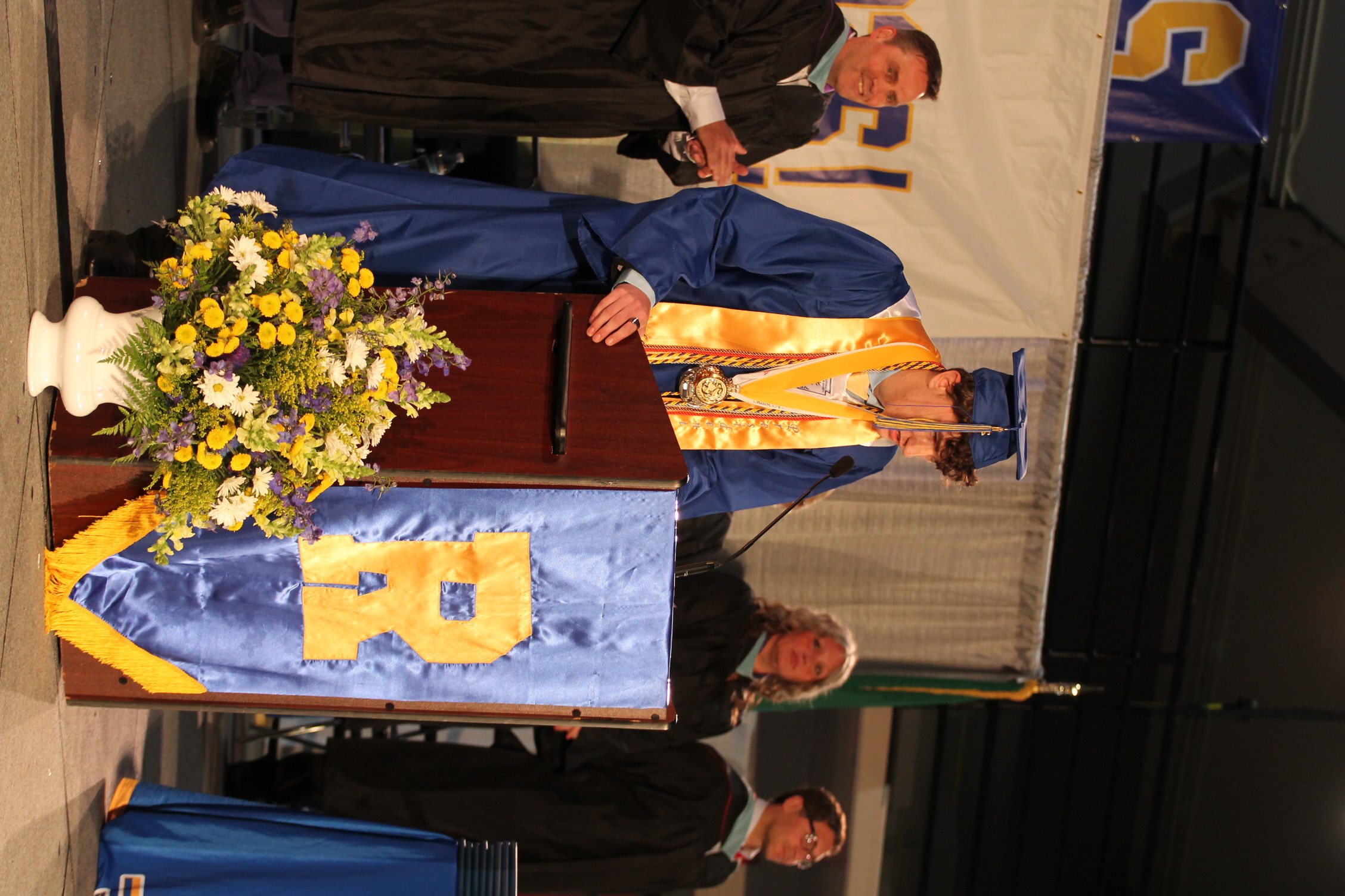 A person in a graduation gown stands at a podium with a microphone. A bouquet of flowers and a banner with the letter R are nearby.