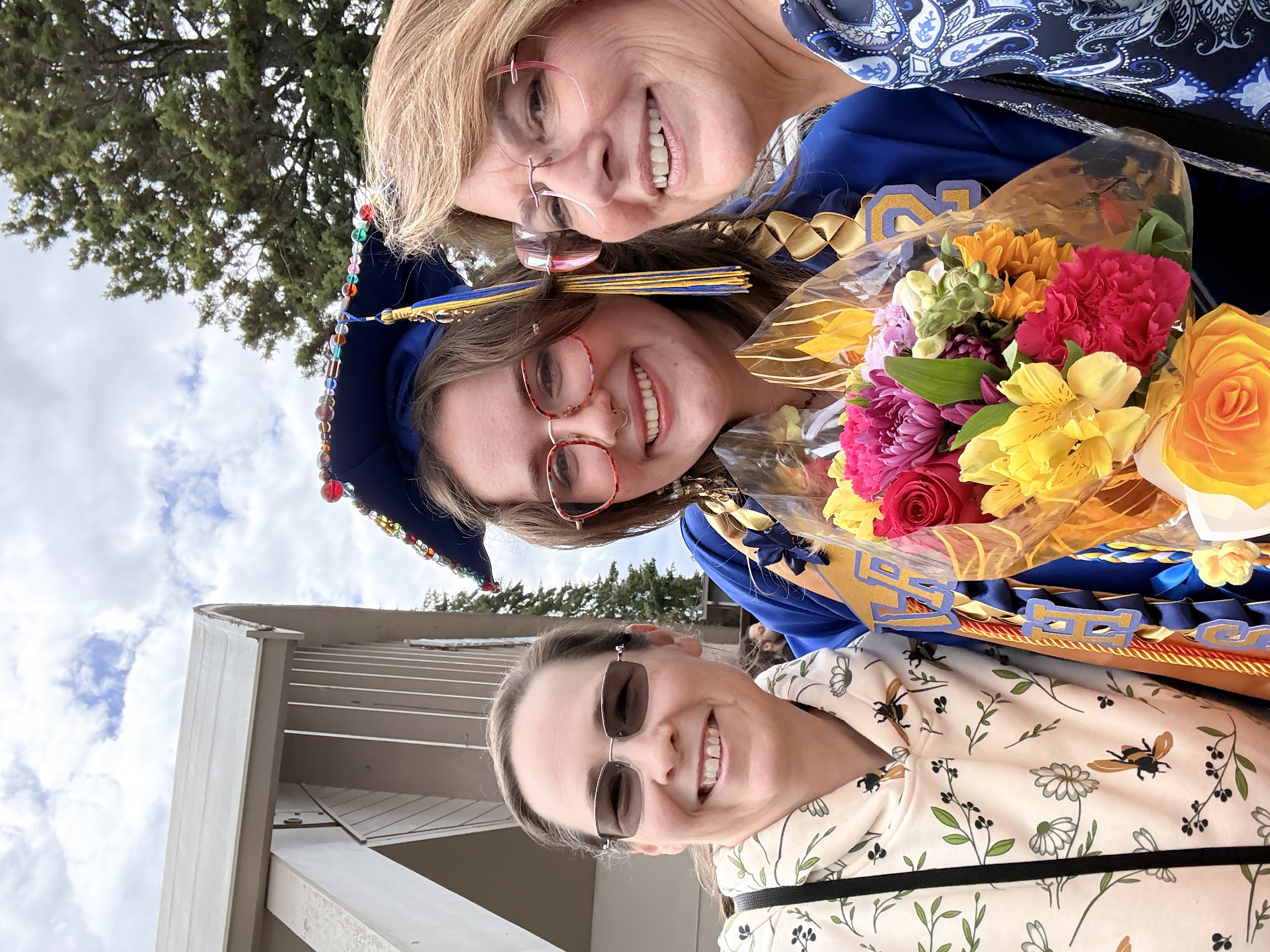 Three women stand together; one in cap and gown, another holds flowers. Background shows a building with trees.