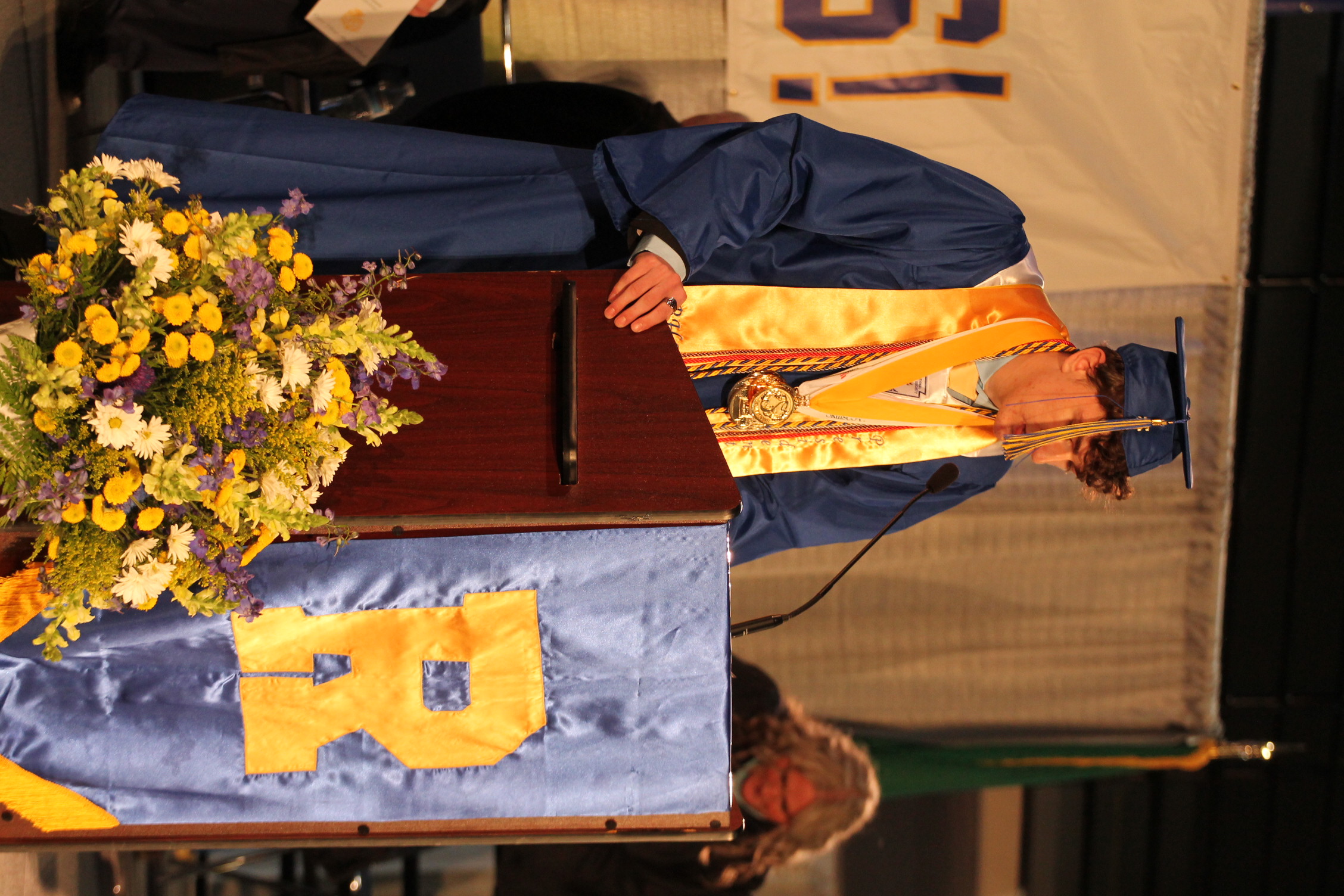 A person in a graduation gown stands at a podium with a microphone, a bouquet of flowers, and a banner reading "R".
