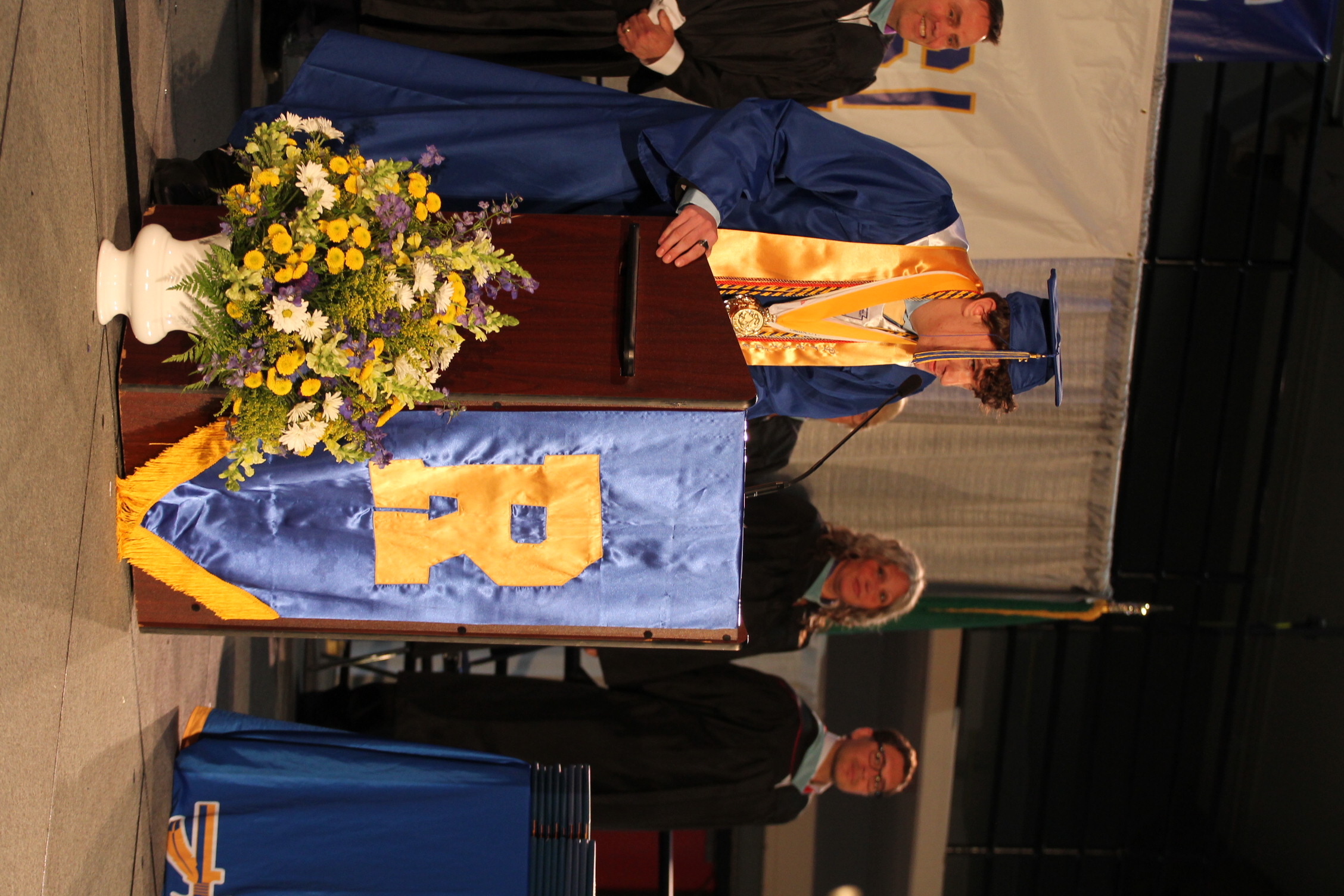 A person wearing a blue graduation gown speaks at a podium. A podium displays the letters "R".