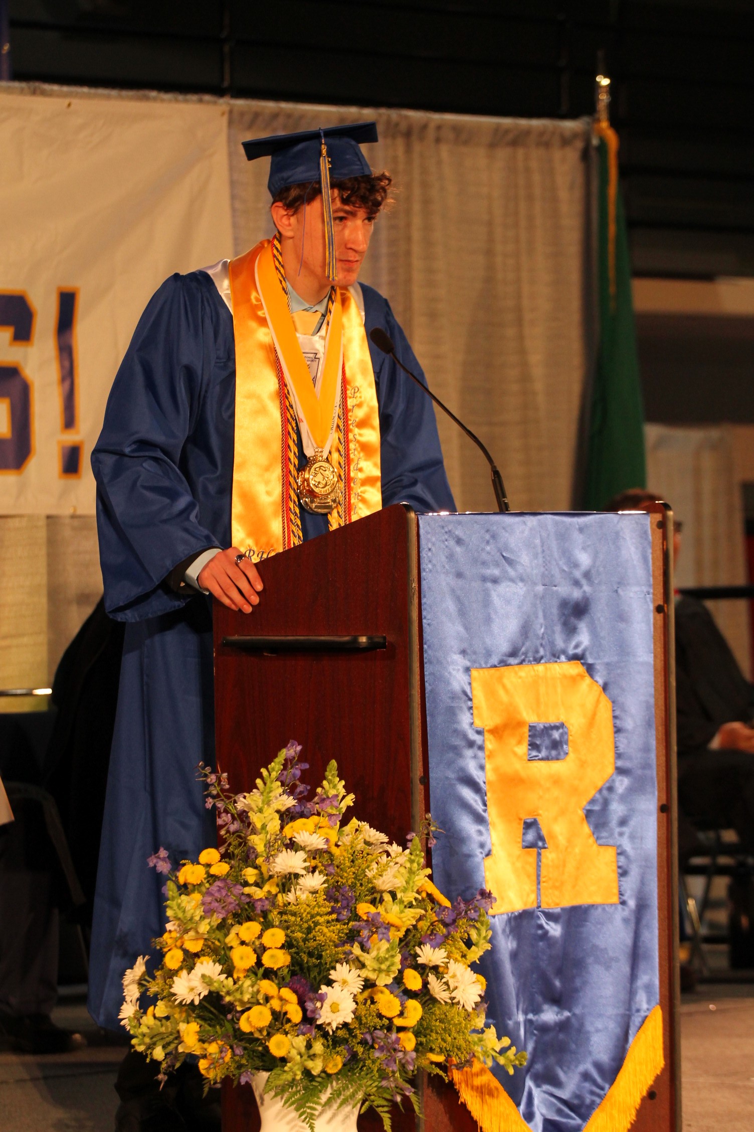 A person in a graduation gown stands at a podium with a banner reading "R" in yellow.