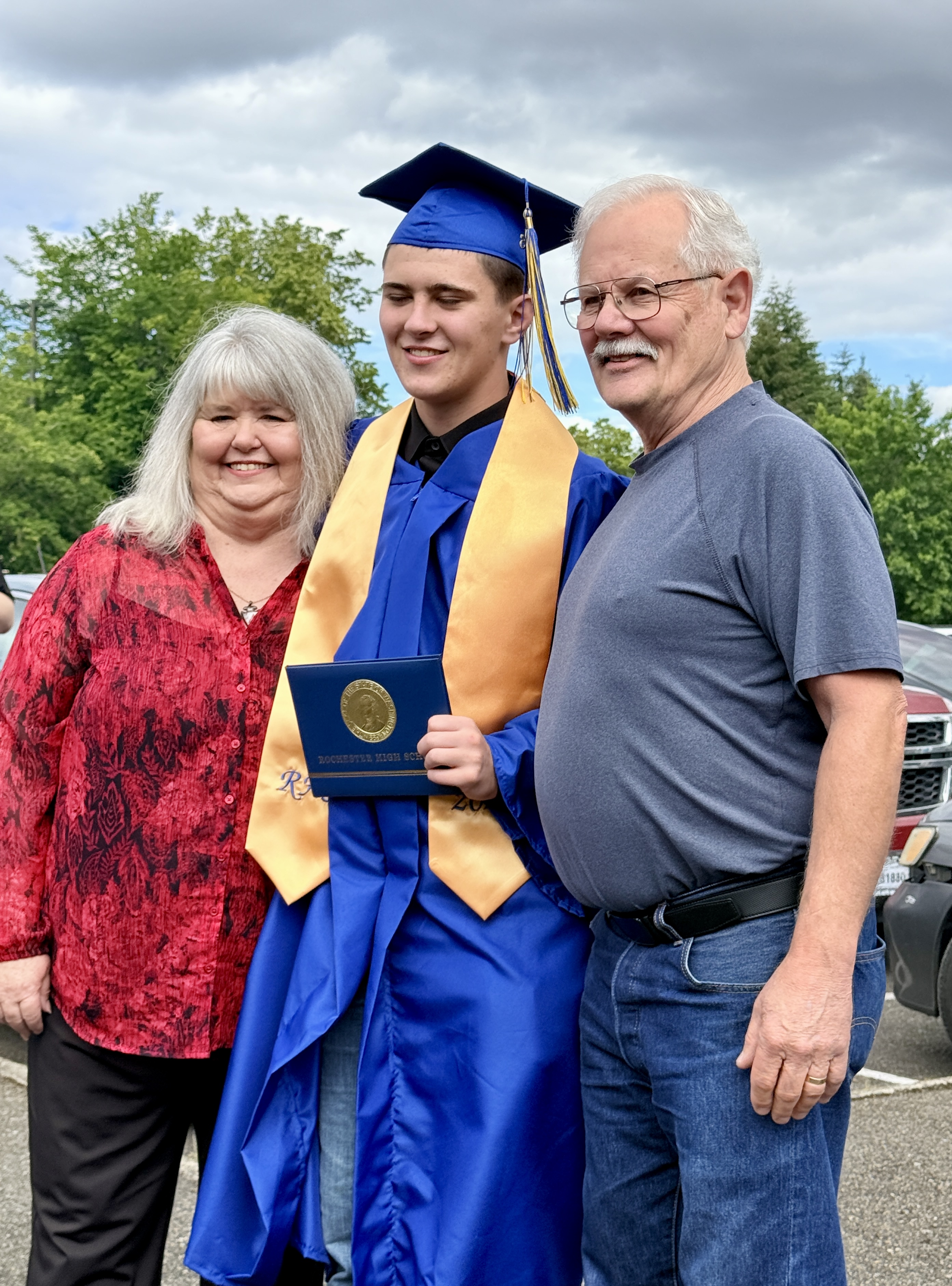 A young man in a graduation gown stands between two adults, holding a diploma. Background includes trees and parked cars.
