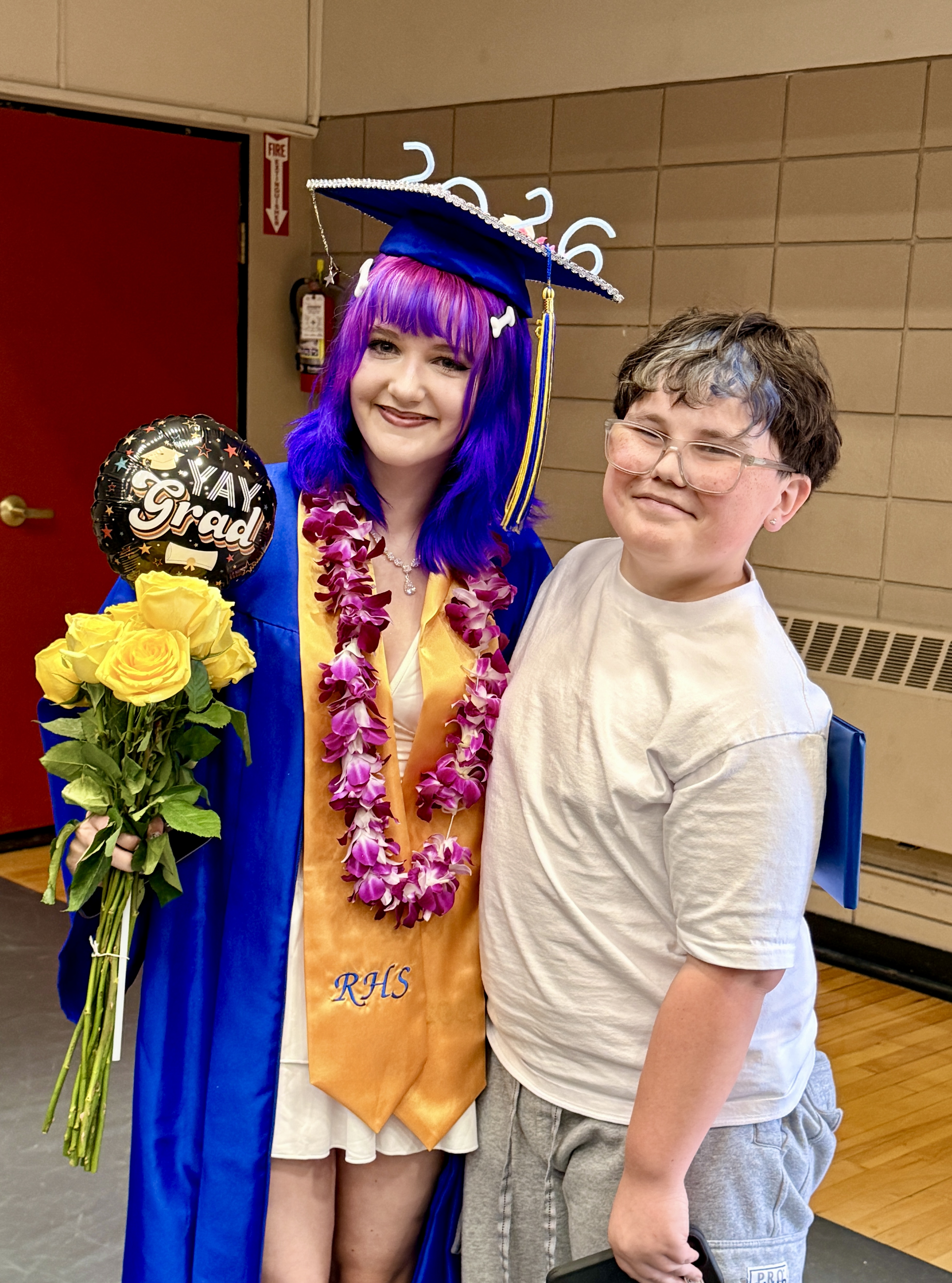 A person with purple hair and a blue gown stands next to a person wearing glasses and a white shirt.