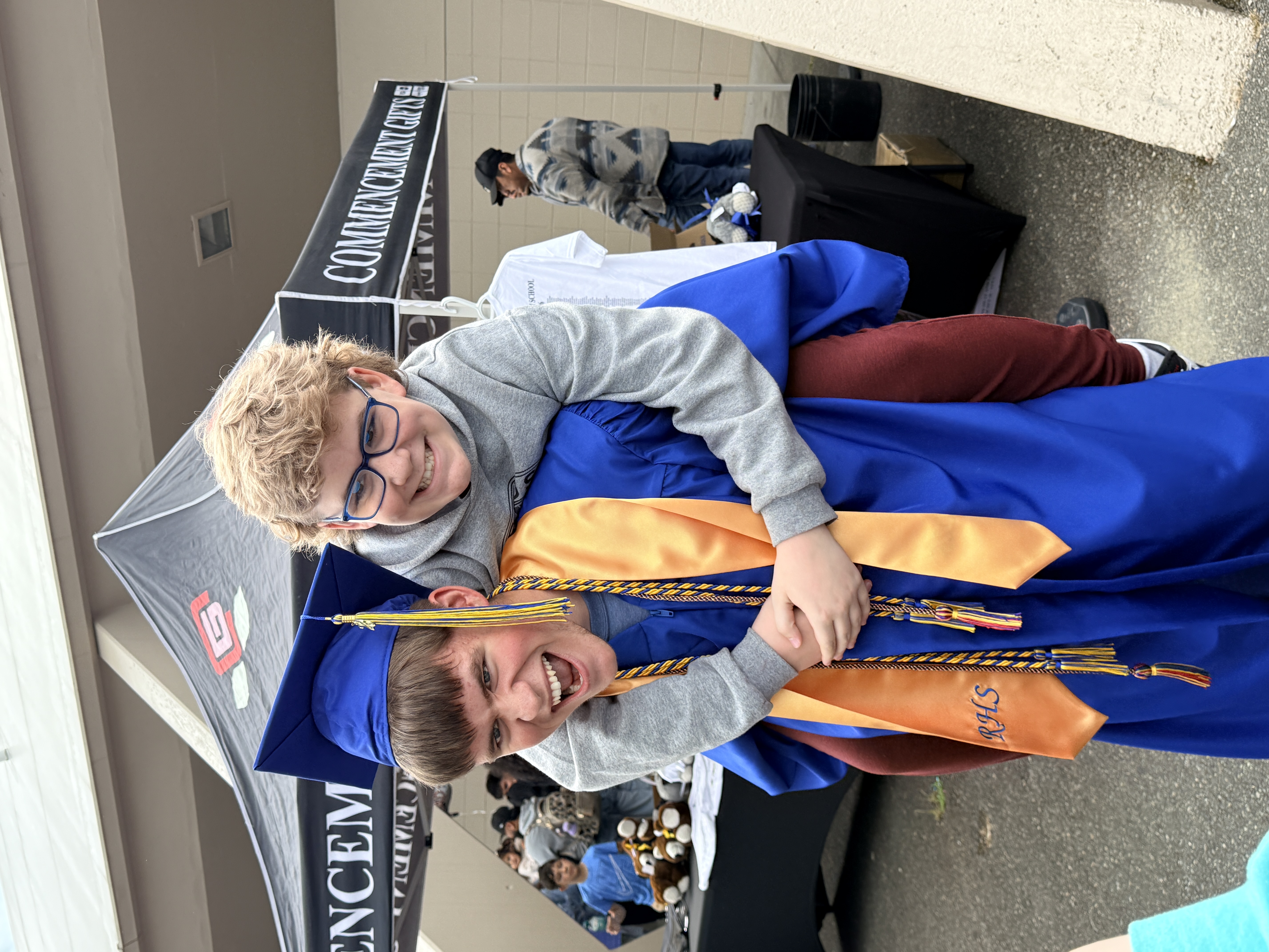 Two boys celebrate, one in a blue graduation gown and the other in a gray shirt, both smiling.