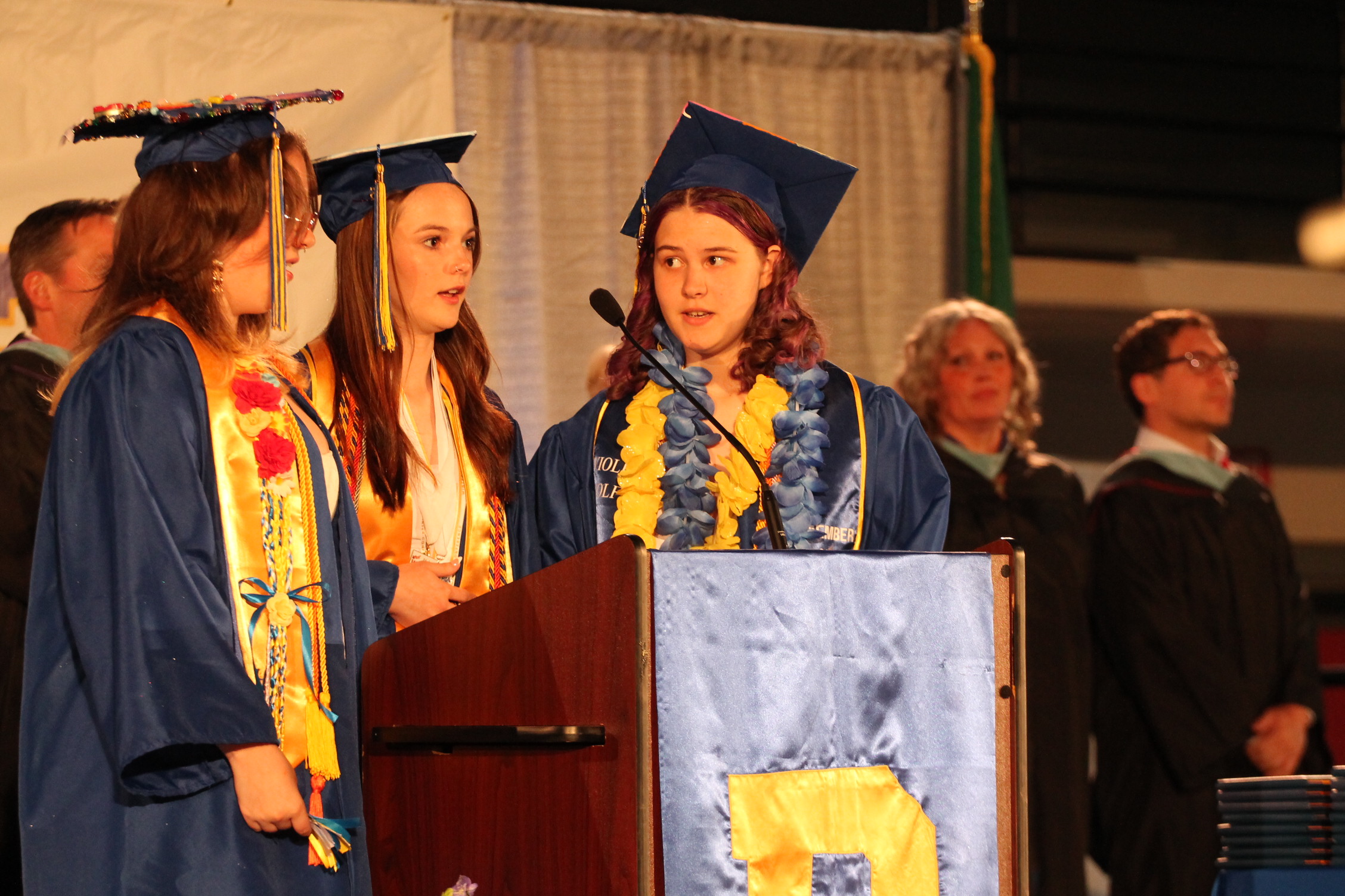 Three women in caps and gowns stand at a podium with a microphone. Behind them, other people, possibly family, stand and watch.