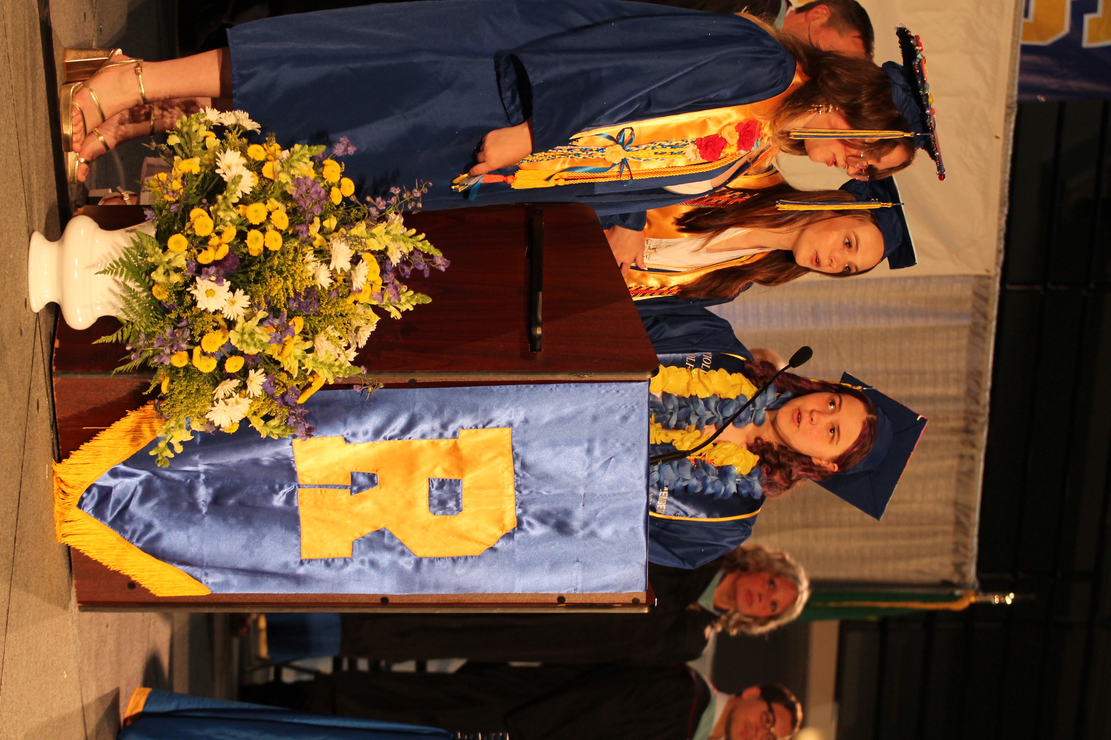 Three women in graduation gowns at a podium with a flower arrangement. The podium has a blue and yellow banner with the letter R.