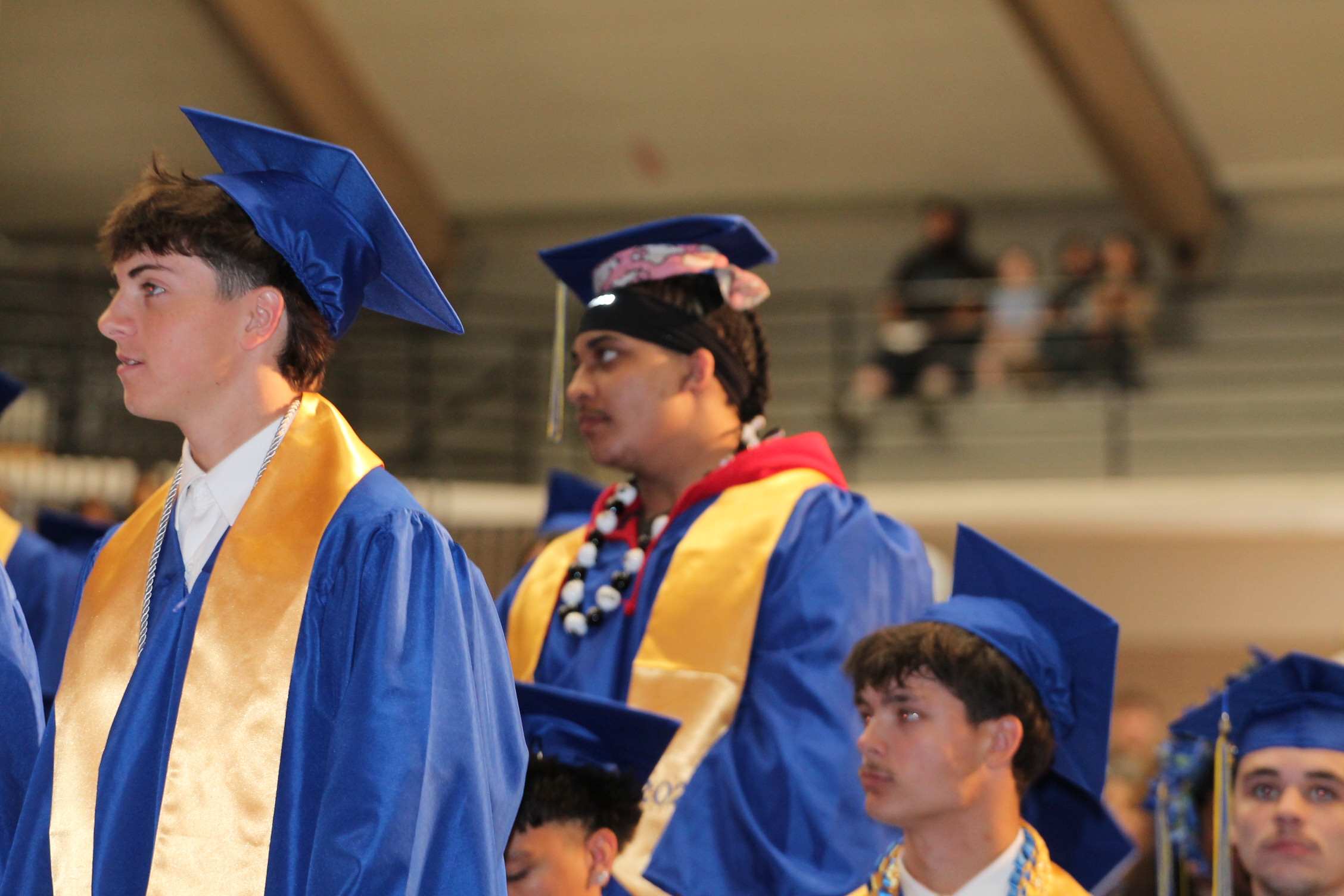 Several people in blue and yellow graduation gowns, with blue caps, stand in a row in a stadium.