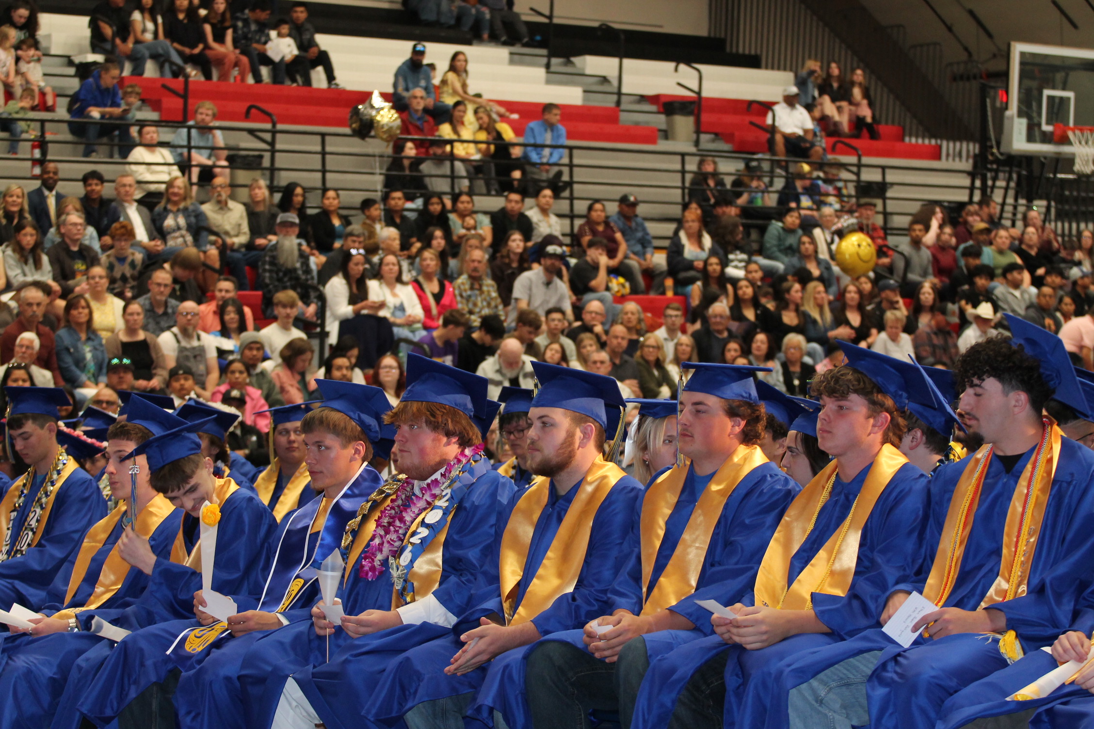 Graduates in caps and gowns seated in rows, looking down, with bleachers full of audience members behind them.