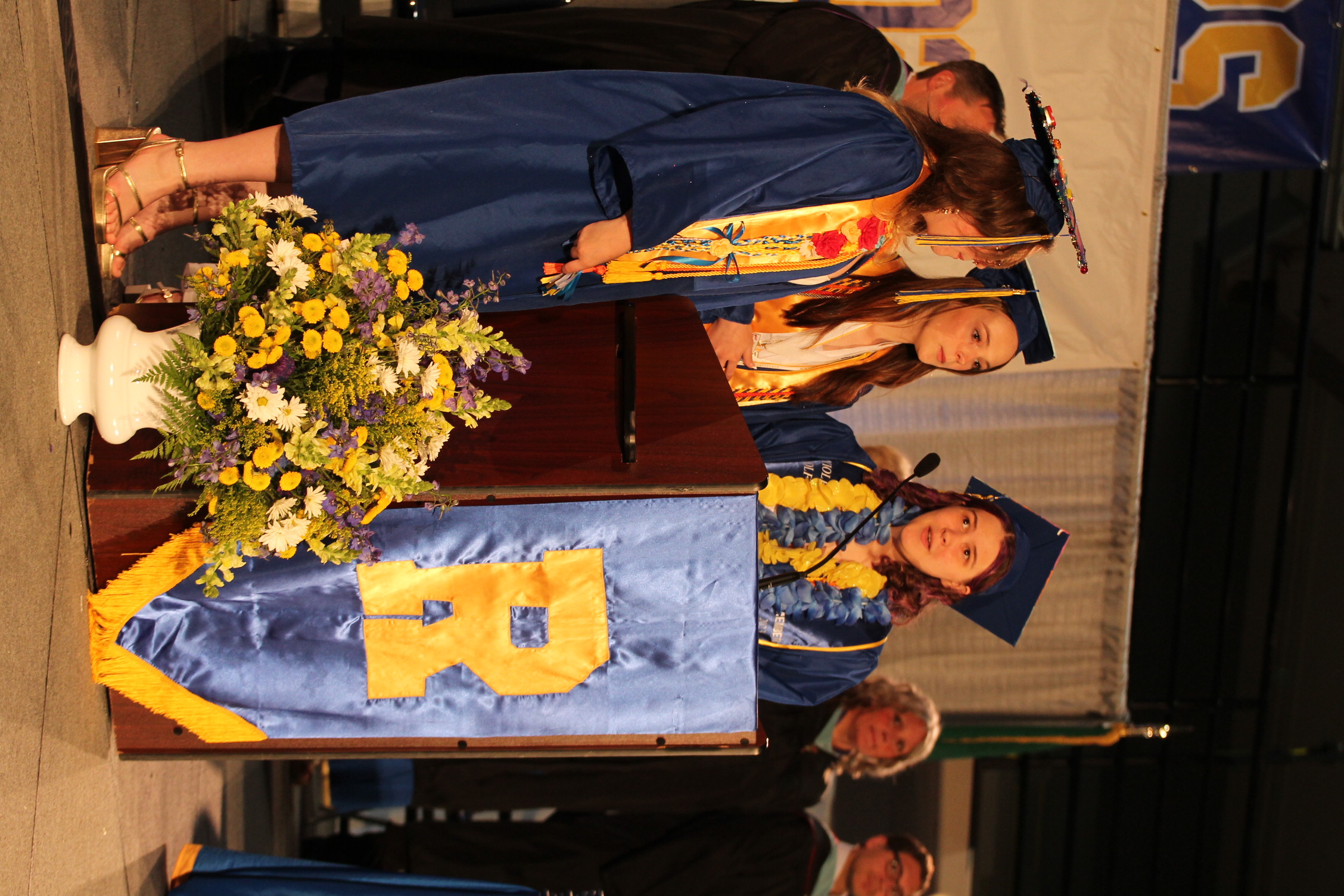 Three women in blue gowns and caps on stage. One speaks at podium with blue banner. Flowers in a vase.