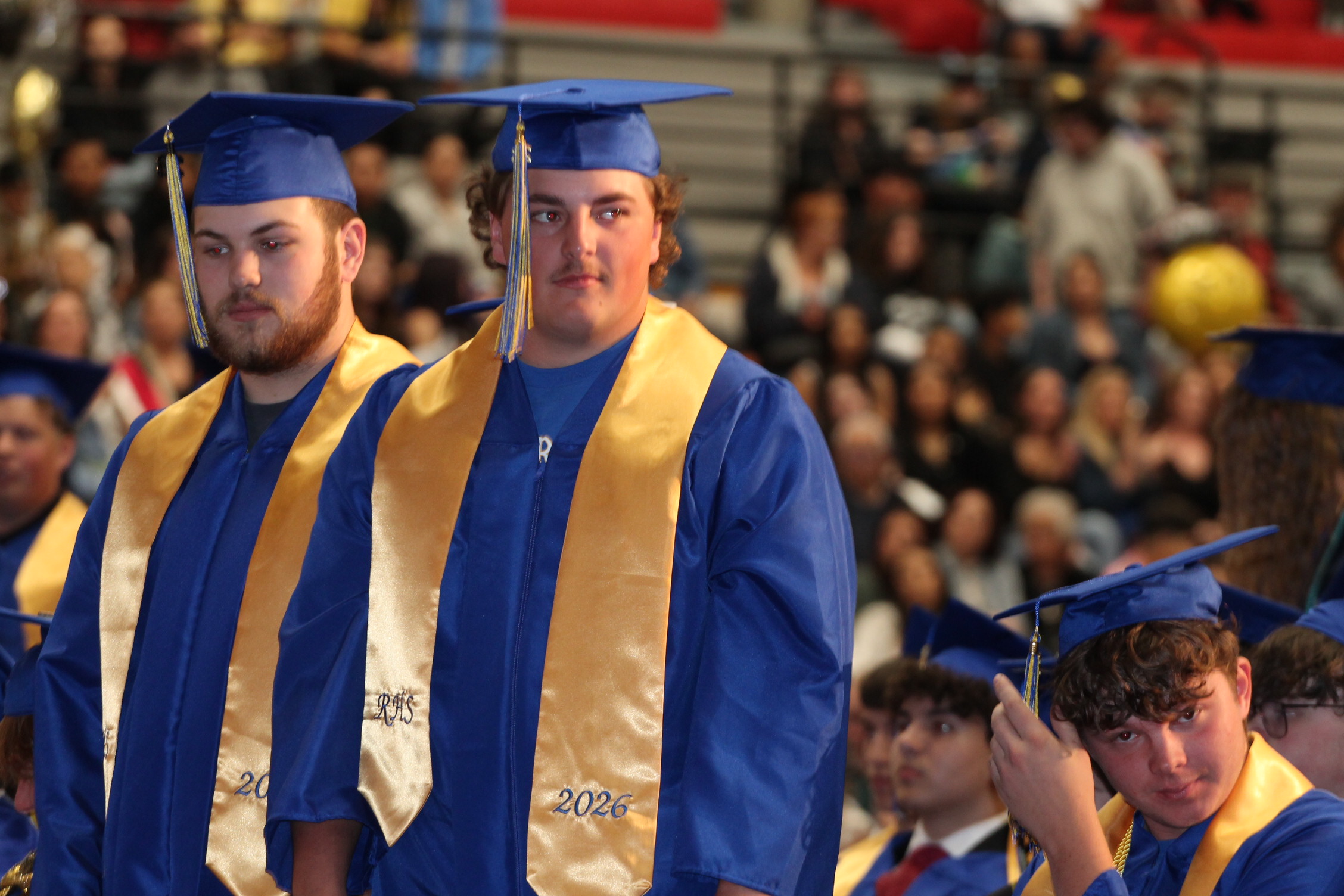 Three individuals in caps and gowns, likely at a graduation ceremony, with a large audience in the background.