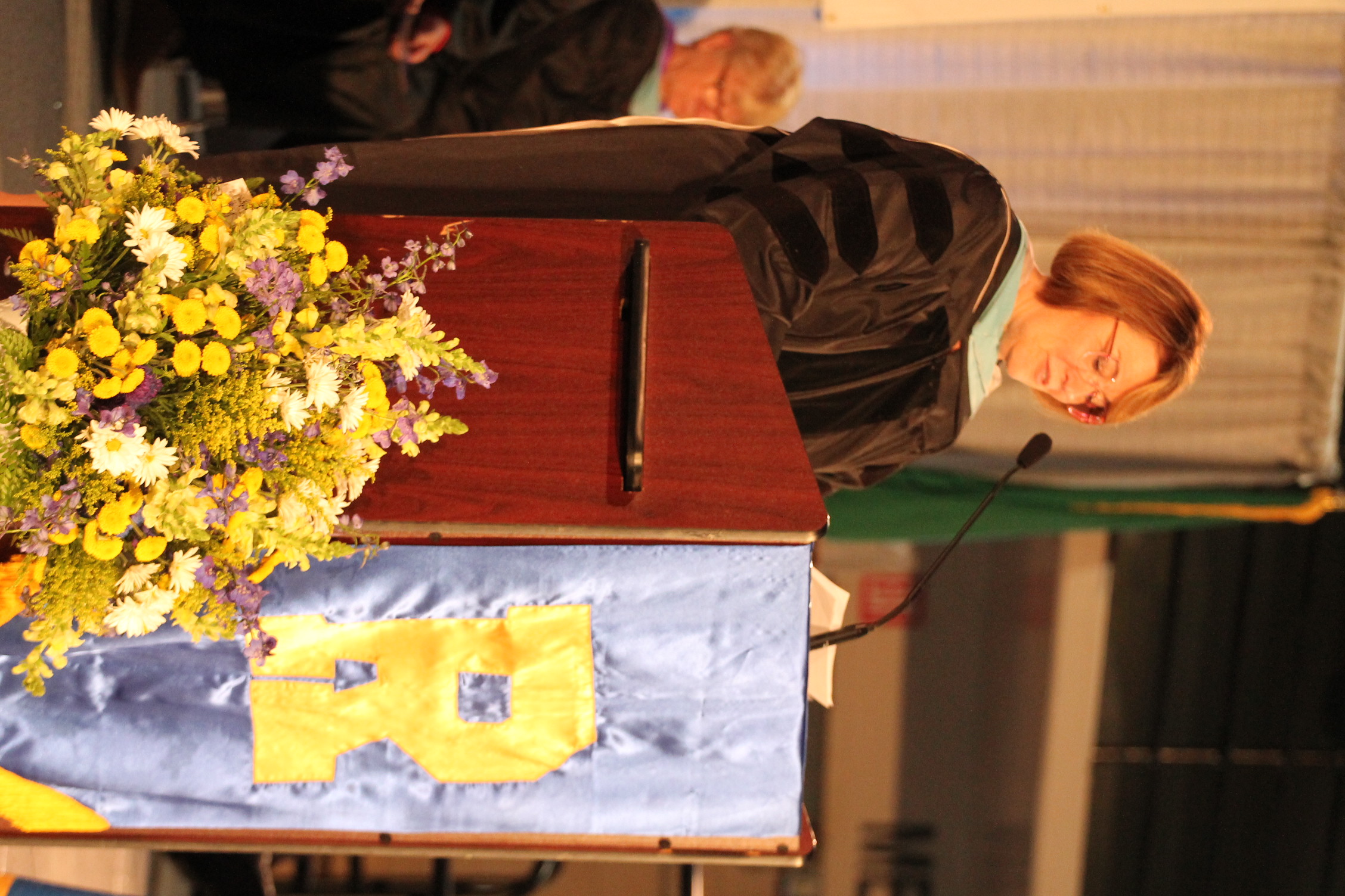 Woman in graduation gown speaks at a podium with a microphone and flowers in front.