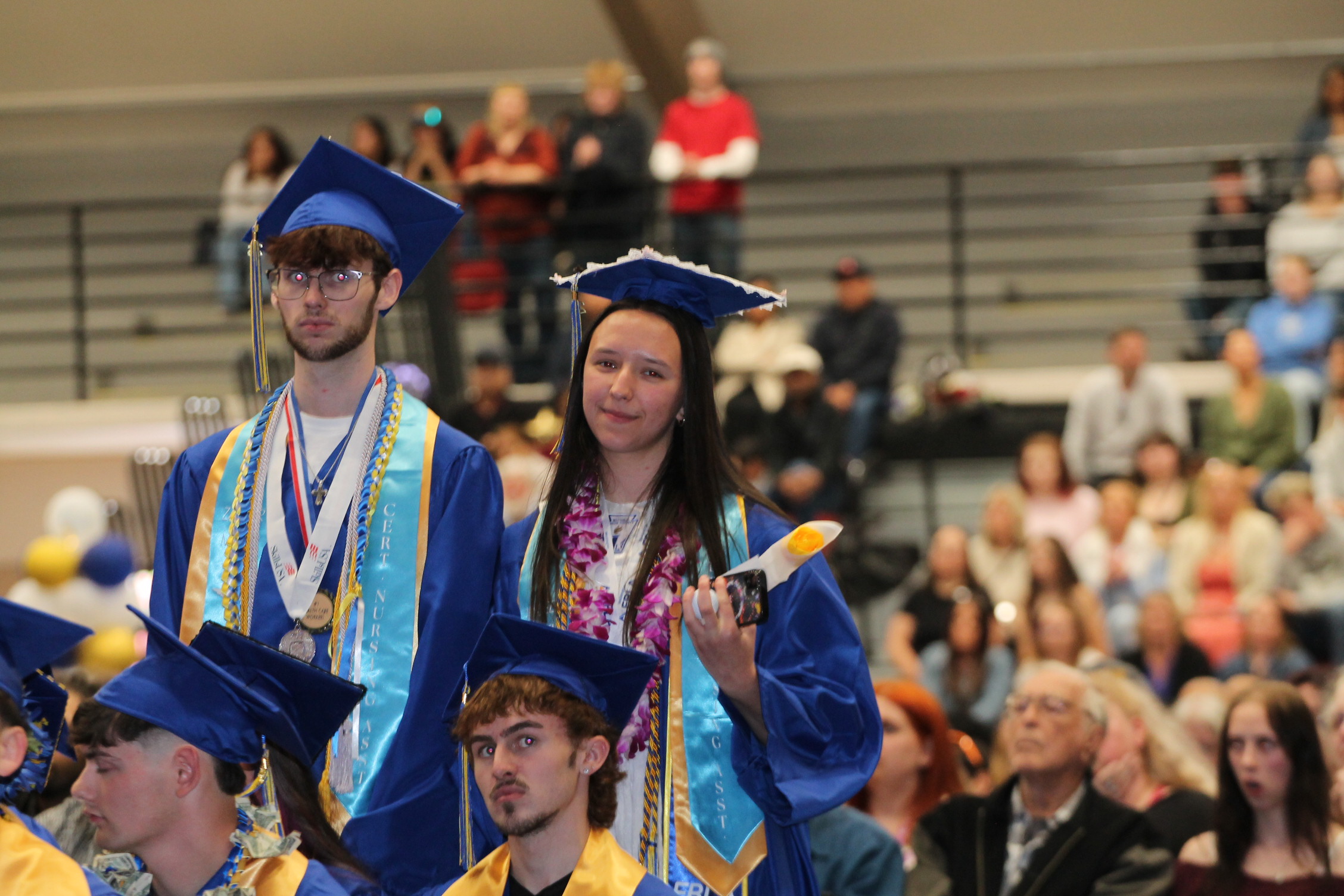 Four individuals in caps and gowns stand in a stadium; a woman holds a diploma. Behind them, spectators sit in bleachers.
