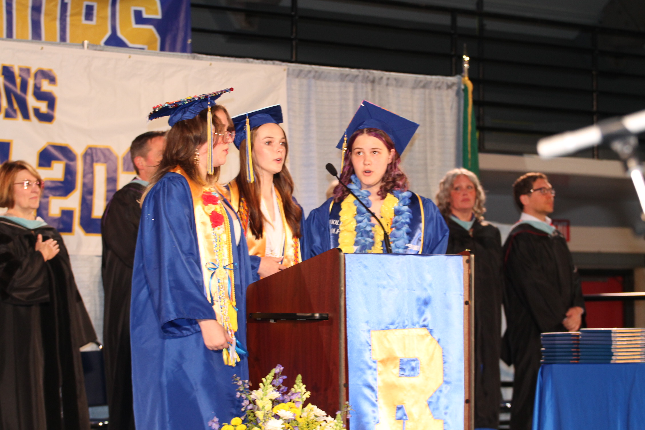 Three women in caps and gowns stand at a podium with a microphone, likely at a graduation ceremony.