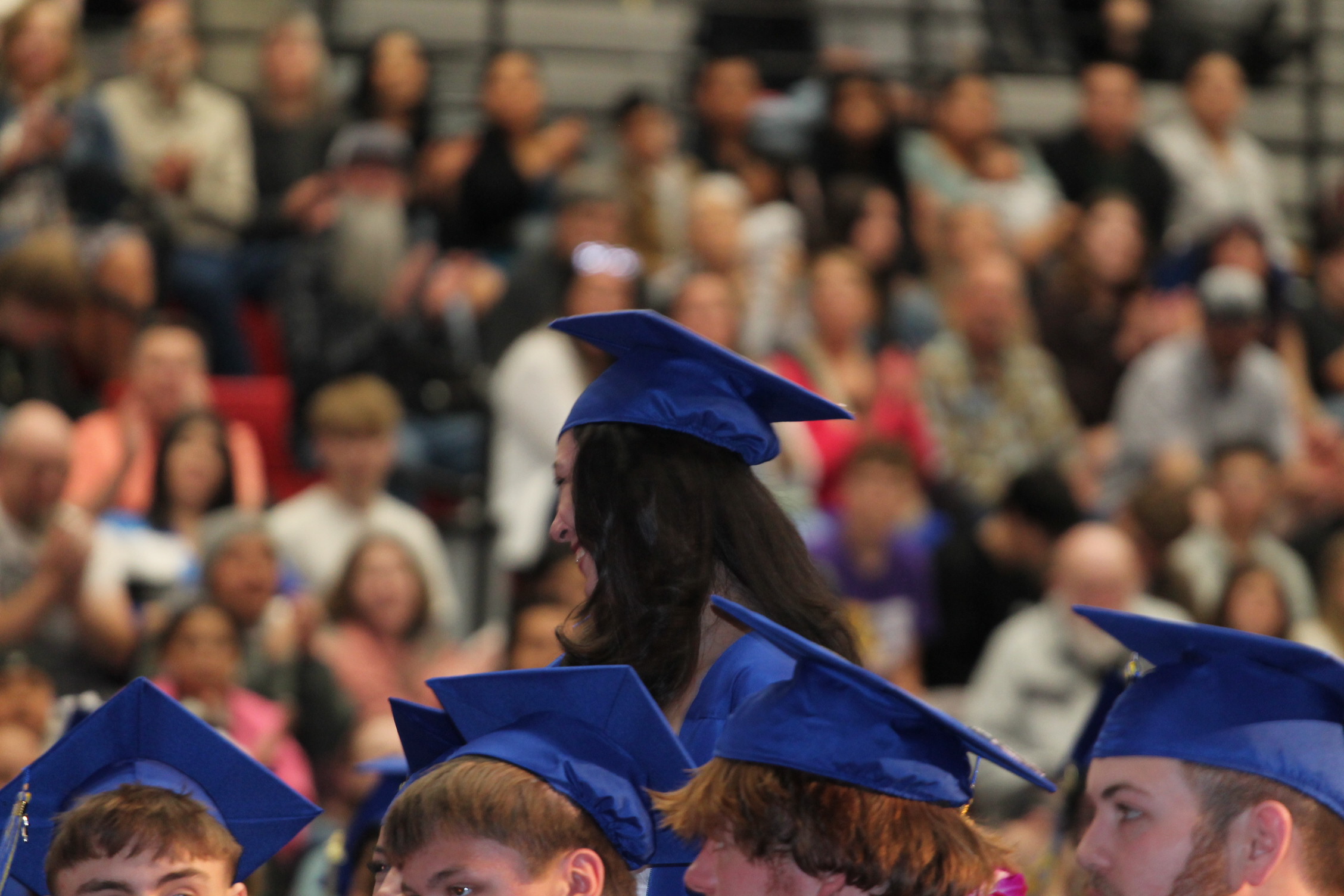 Graduates in blue caps stand in line, facing the audience. The audience, seated in rows, is blurred.