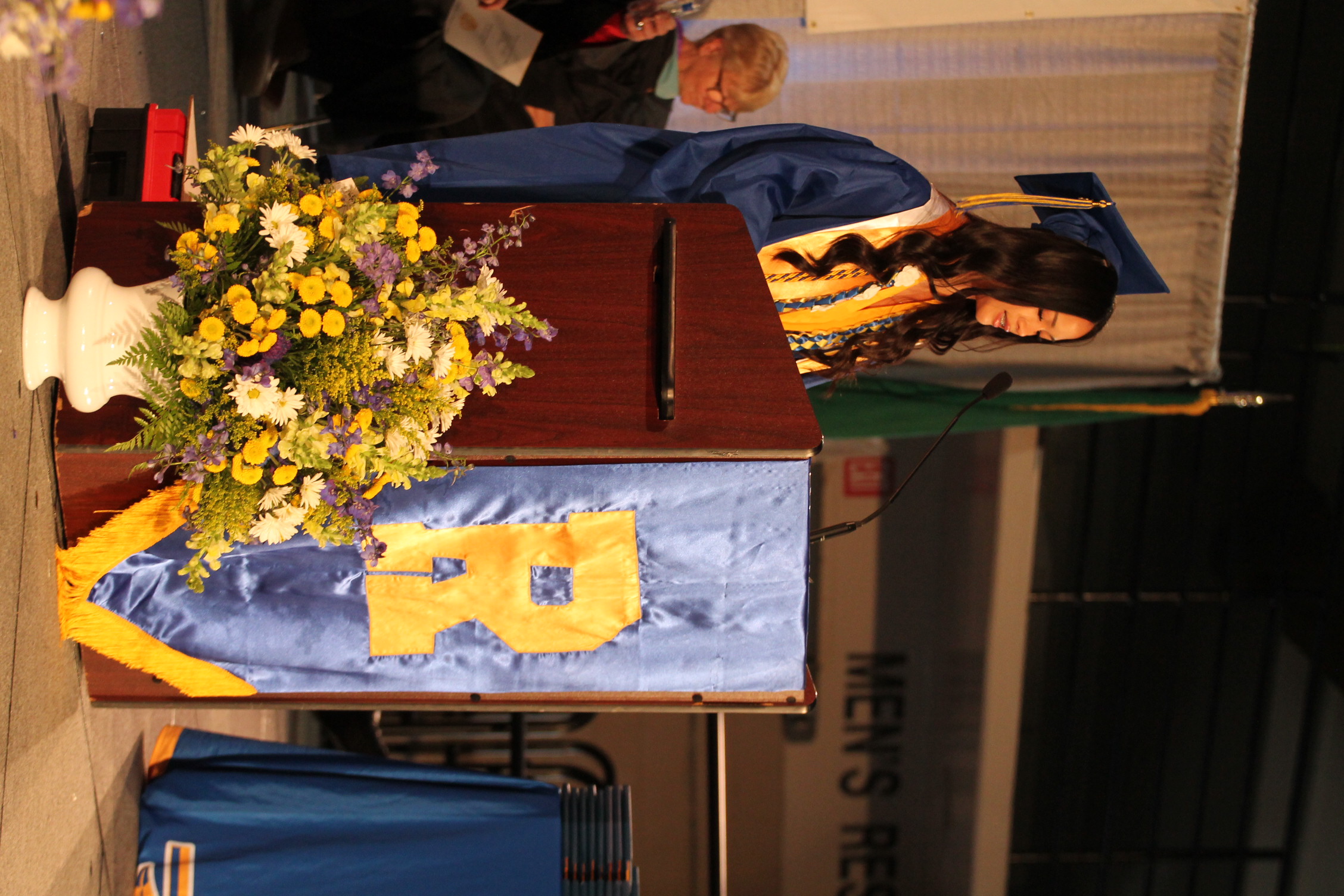 Woman in cap and gown standing at a podium with a microphone, flowers, and a blue banner.