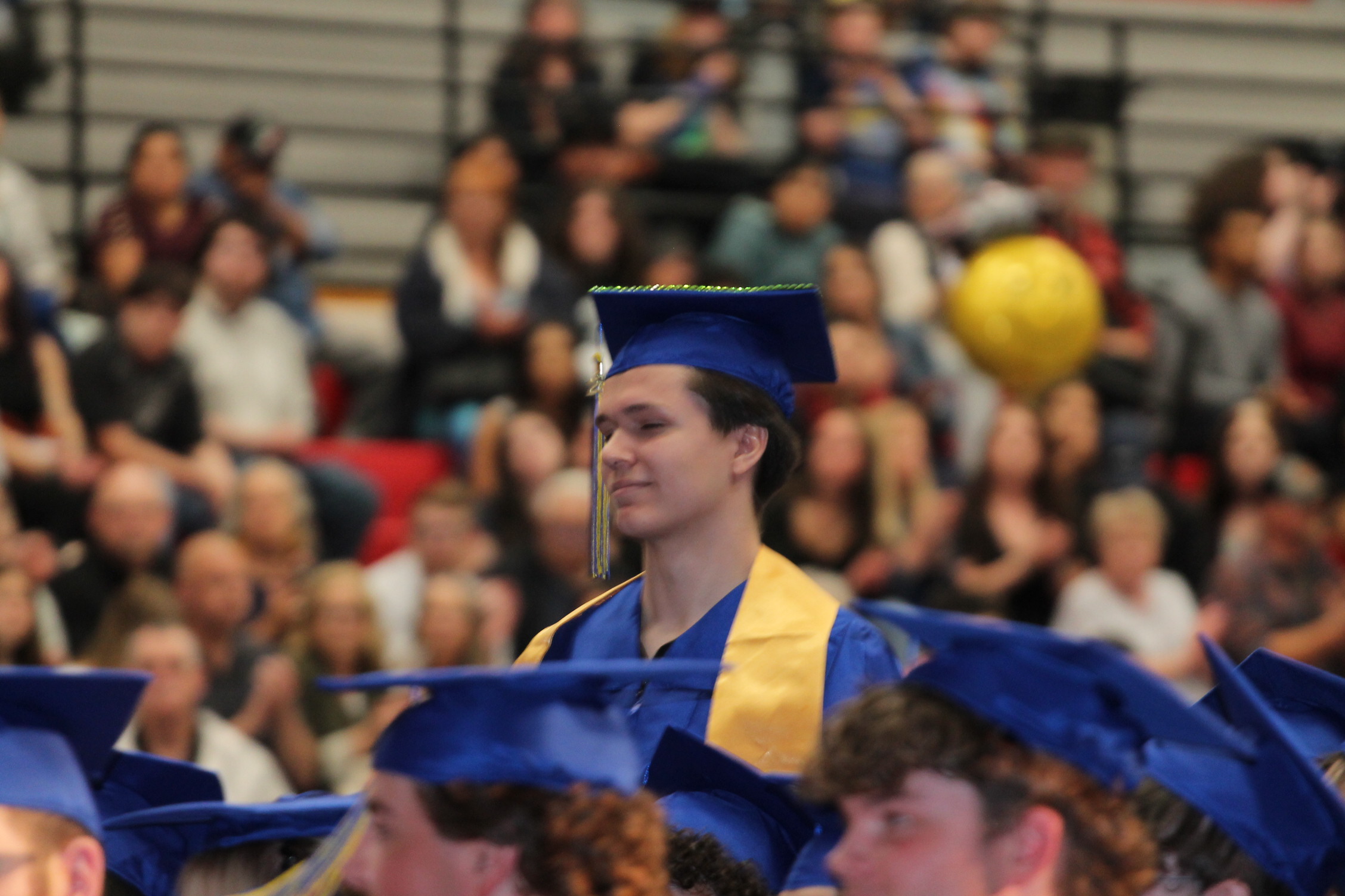 A person in a blue graduation gown stands in front of a crowd wearing similar caps.