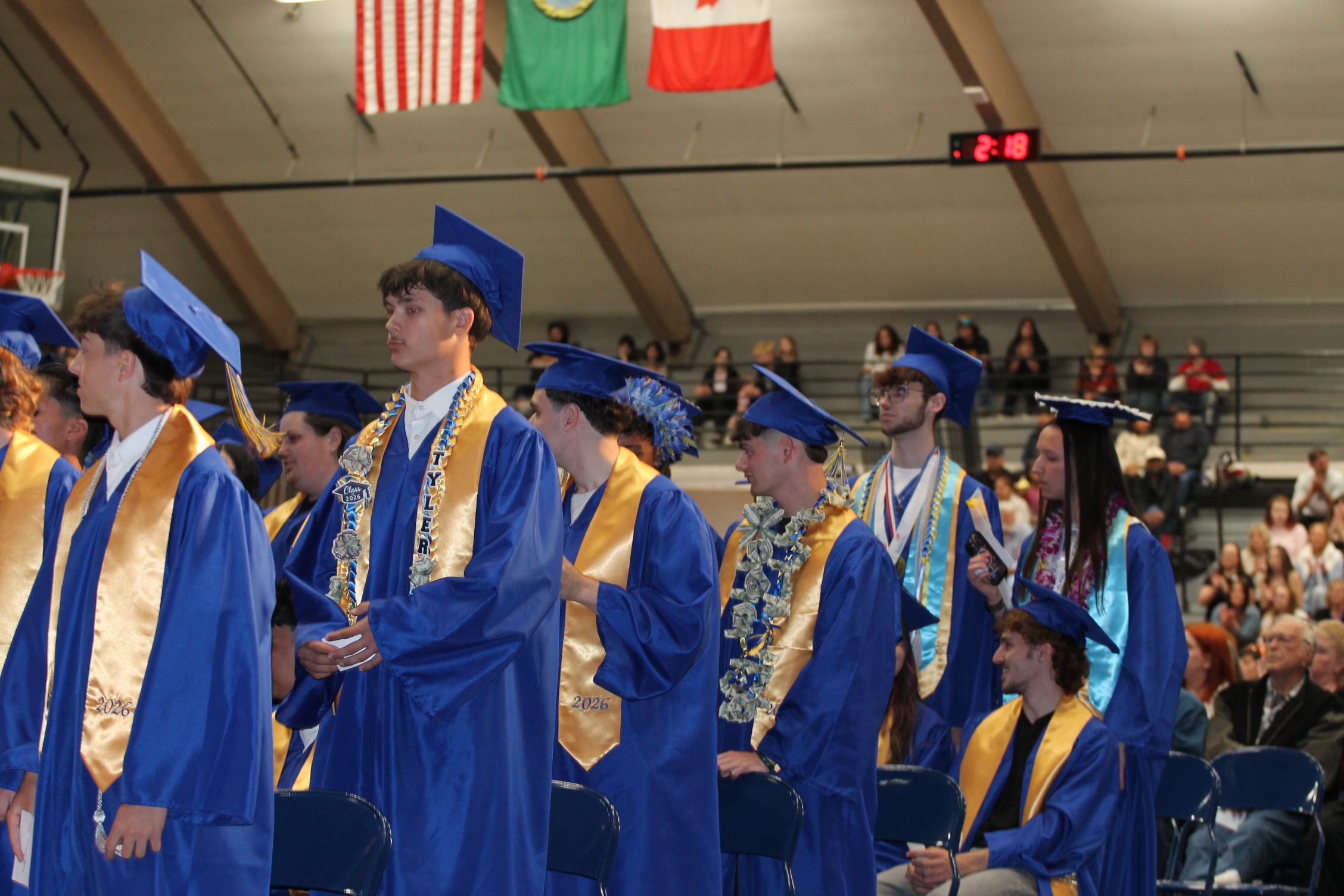 People in blue graduation gowns and hats stand in a line in a gymnasium. Flags hang above.
