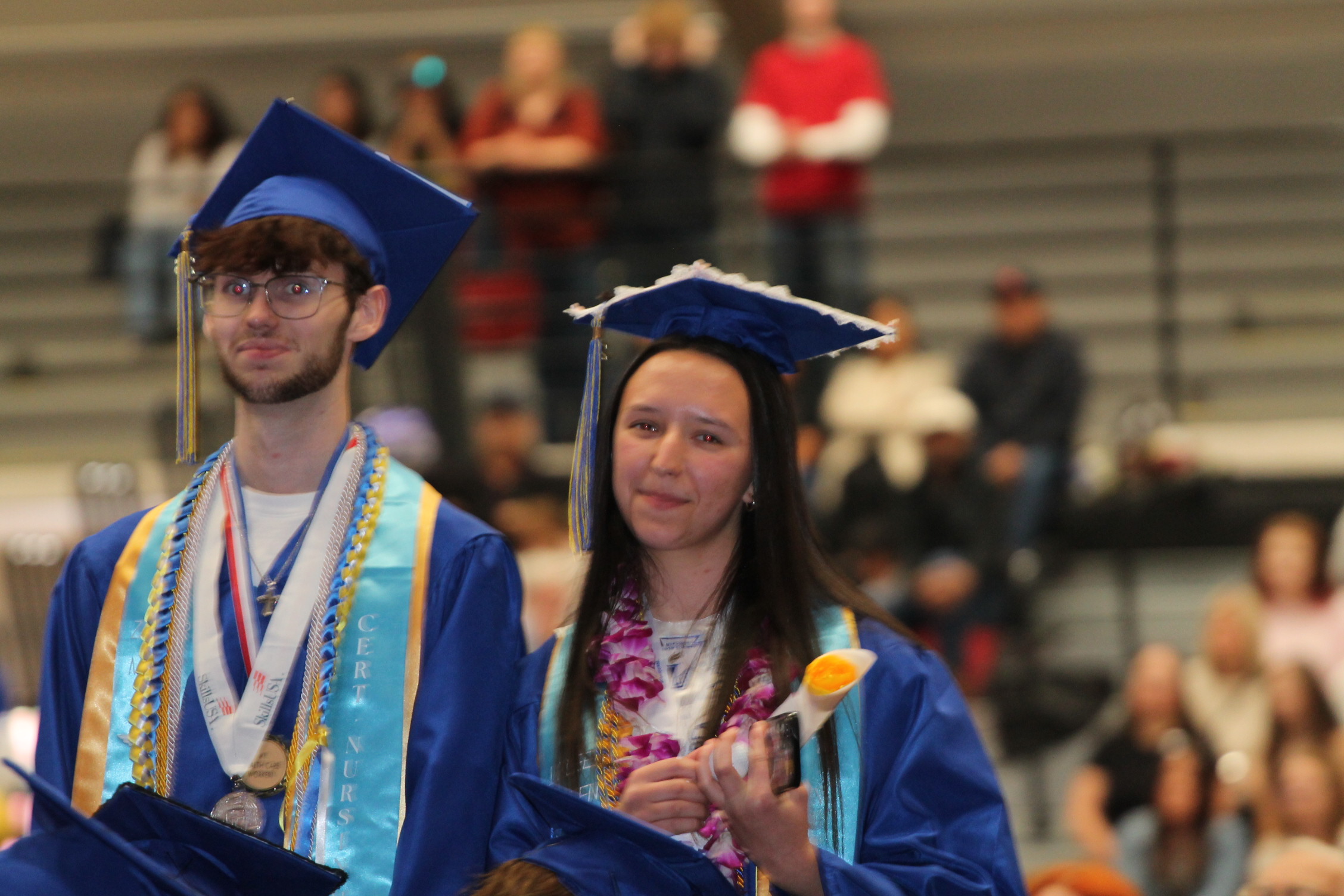 Two graduates in blue gowns and caps stand in a stadium. A man wears glasses, a medal, and a lei. A woman holds a flower.