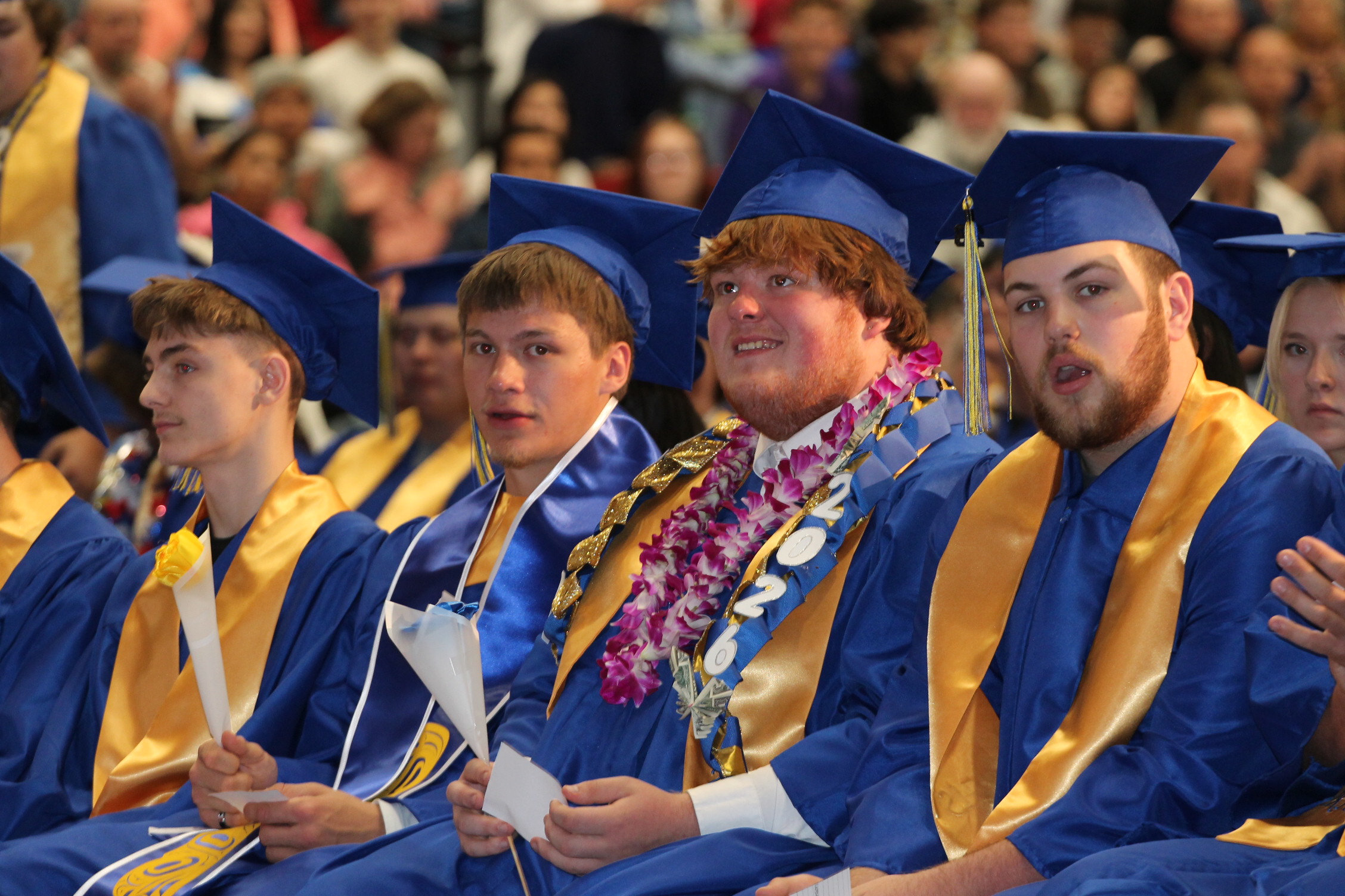 Several young adults in blue and gold gowns, some wearing leis, sit among a crowd.