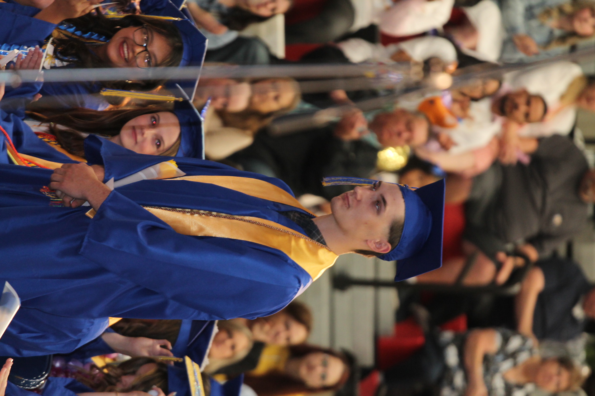 A male student in a blue gown stands in front of seated people, likely during a graduation ceremony.