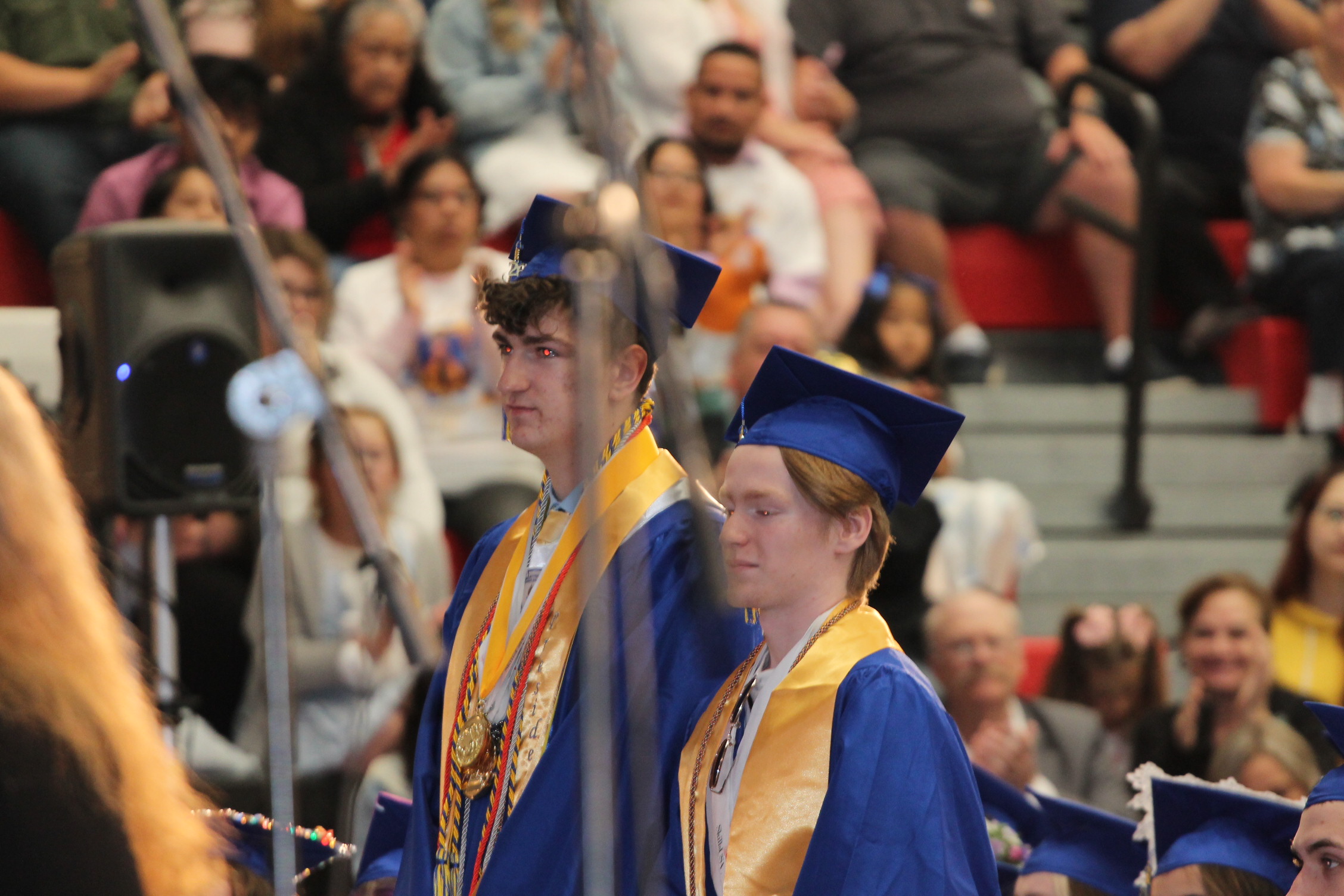 Two students in blue and gold gowns and caps stand in front of a seated audience at an event.