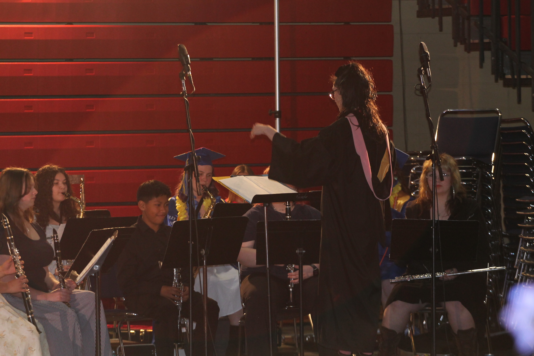 People in graduation robes sit and stand on a stage, playing instruments with a director leading them.
