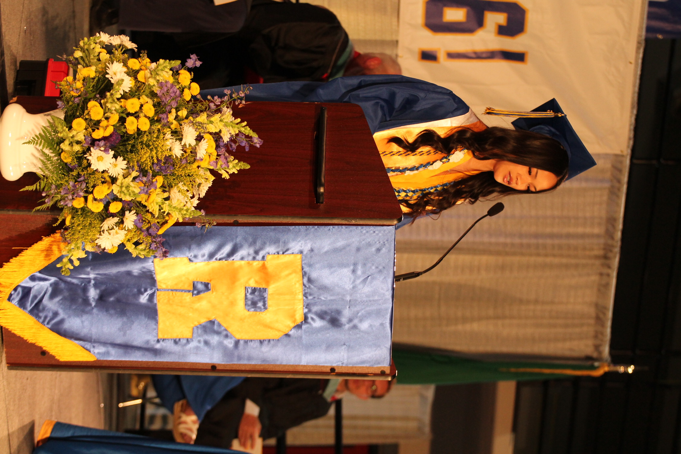 A woman in a graduation gown and cap speaks at a podium with a microphone. Behind her, people sit.