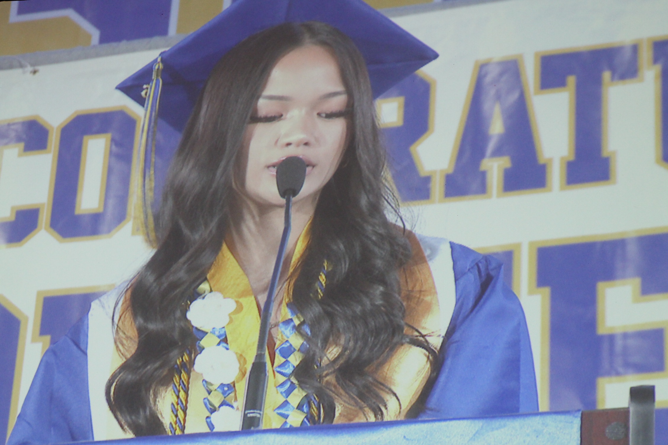 A person in a blue gown with a blue hat speaks into a microphone at a ceremony.