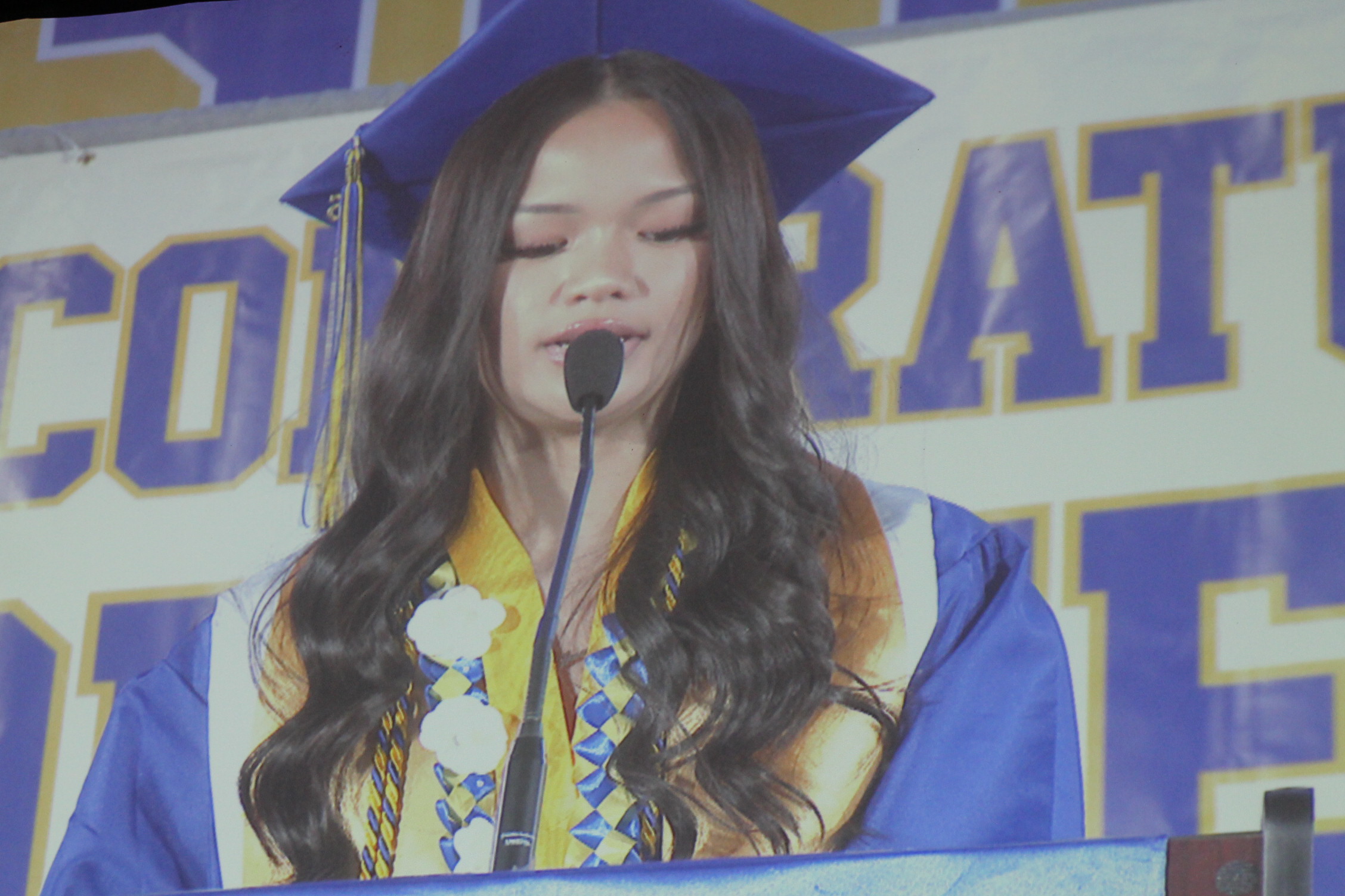 Person in a blue cap and gown, addressing a crowd, speaking into a microphone, with a backdrop reading "CONGRATULATIONS".