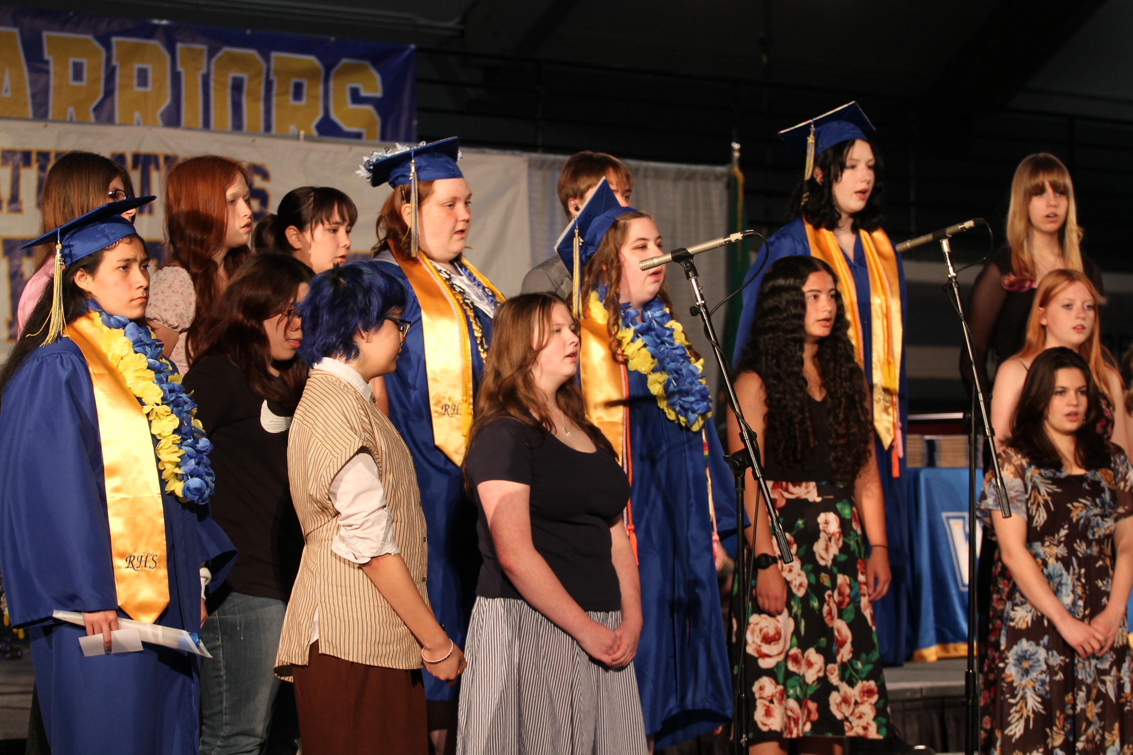 People on a stage wearing graduation attire, with some holding microphones. A banner behind them reads "Grads".