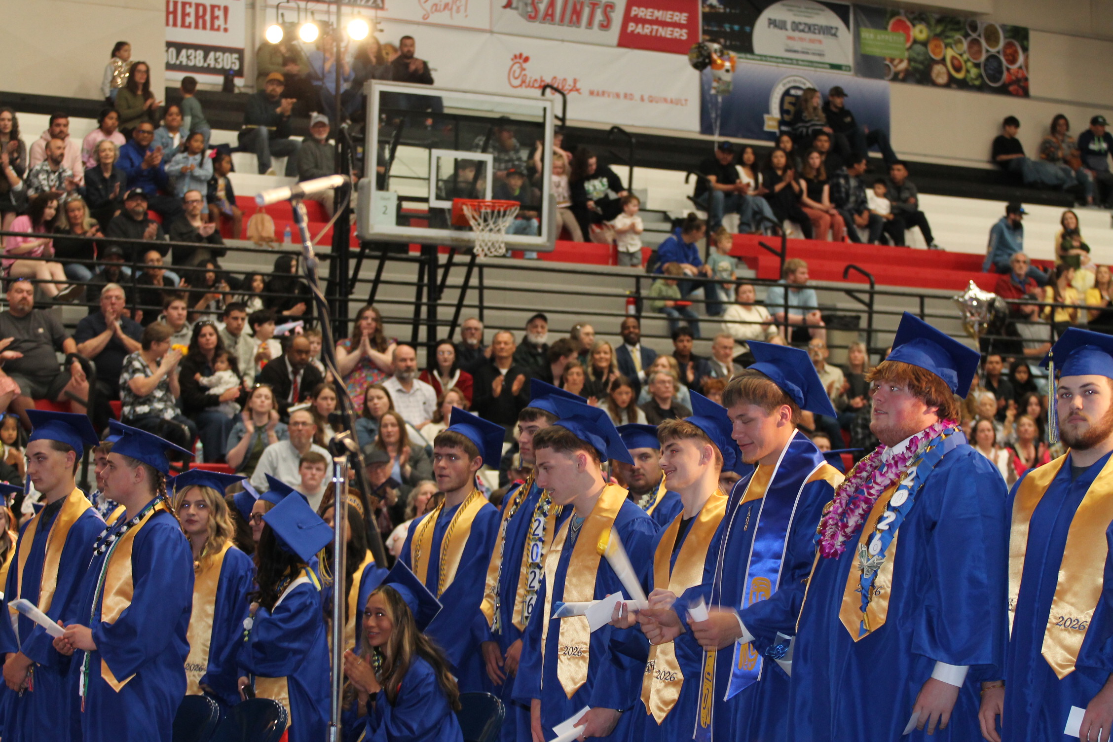Graduates in blue gowns and caps stand in line. Audience seated in bleachers behind them.