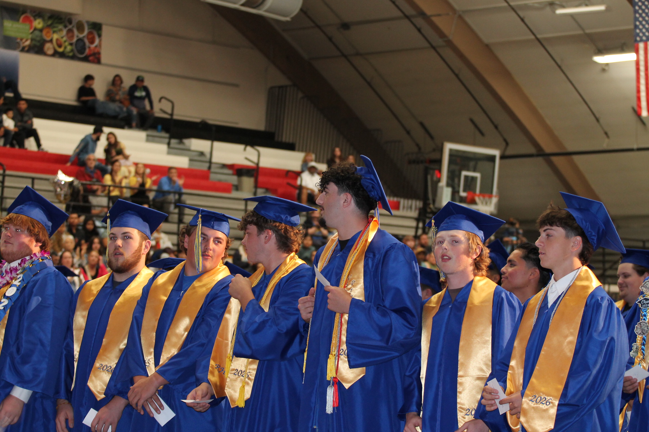 Graduates in blue gowns with gold sashes and caps stand in rows, facing an audience in a gym.