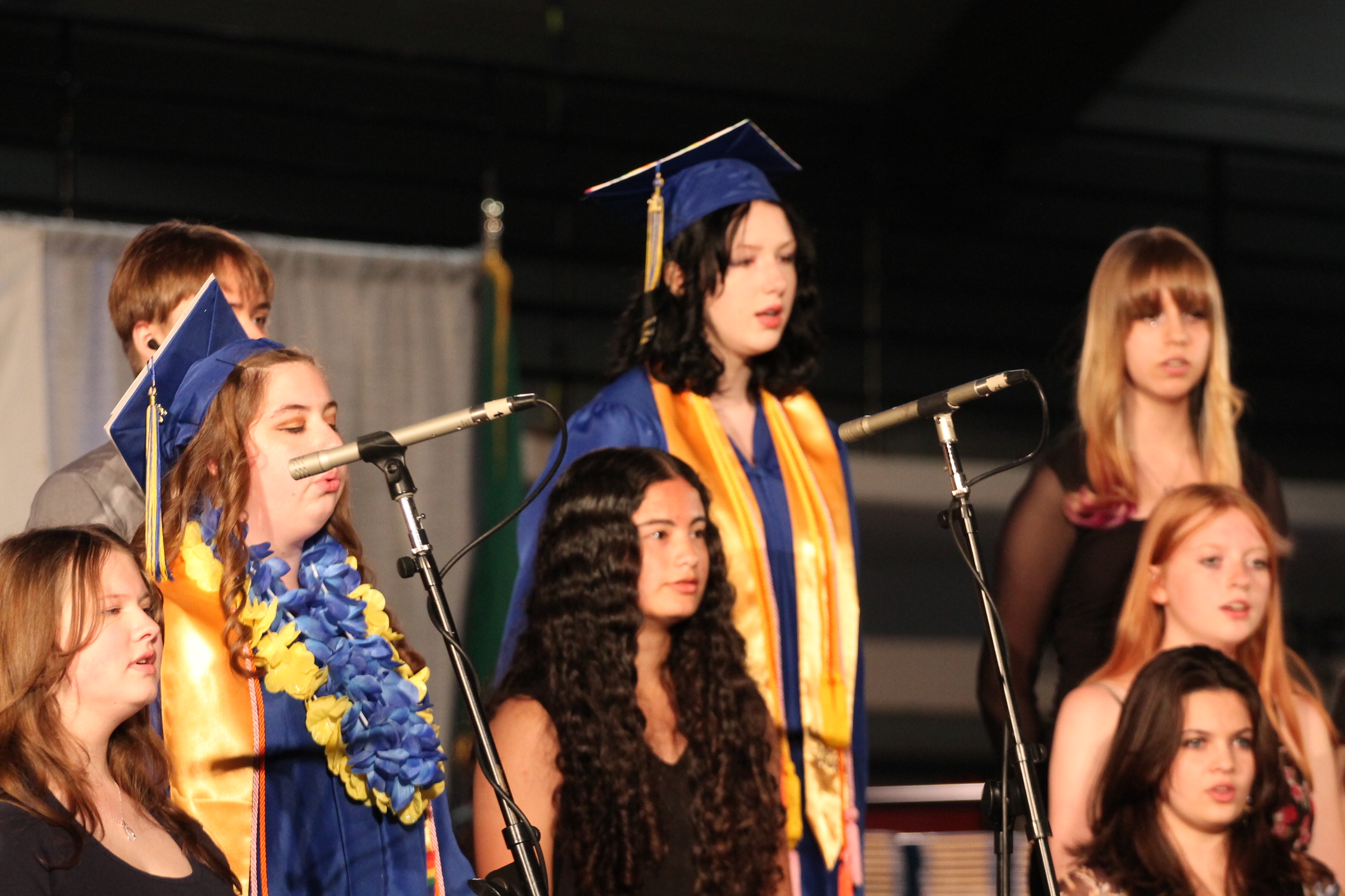 Several young women in blue graduation gowns with yellow sashes stand on stage with microphones.