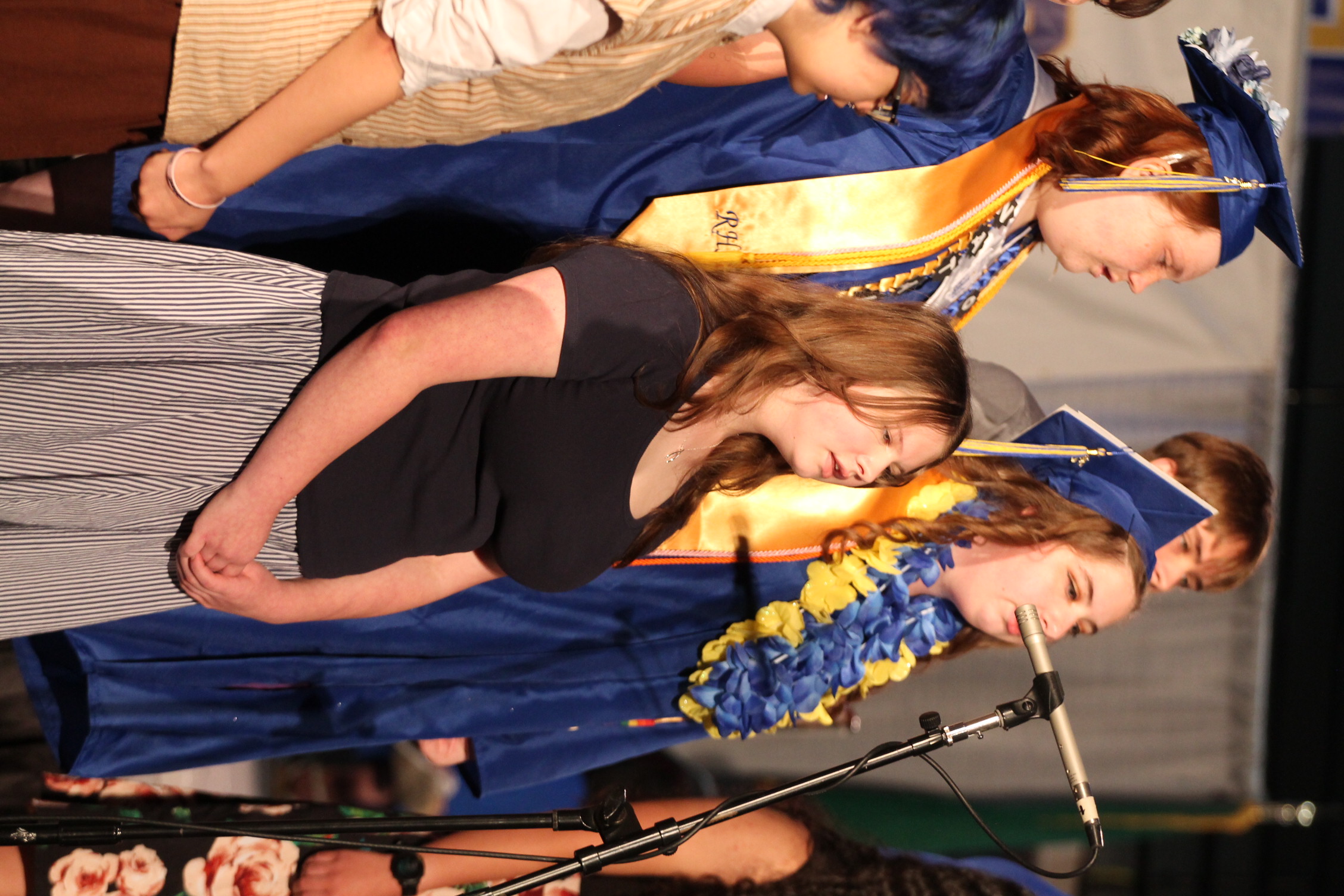 Group of people on stage; three in blue gowns with gold sashes, one in a black shirt, singing.