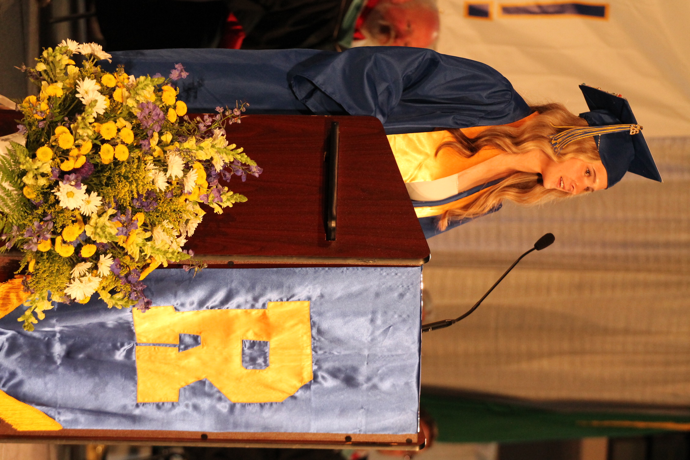 Woman in cap and gown standing at a podium, microphone in front, yellow flowers on the table, and banner with the letter R.