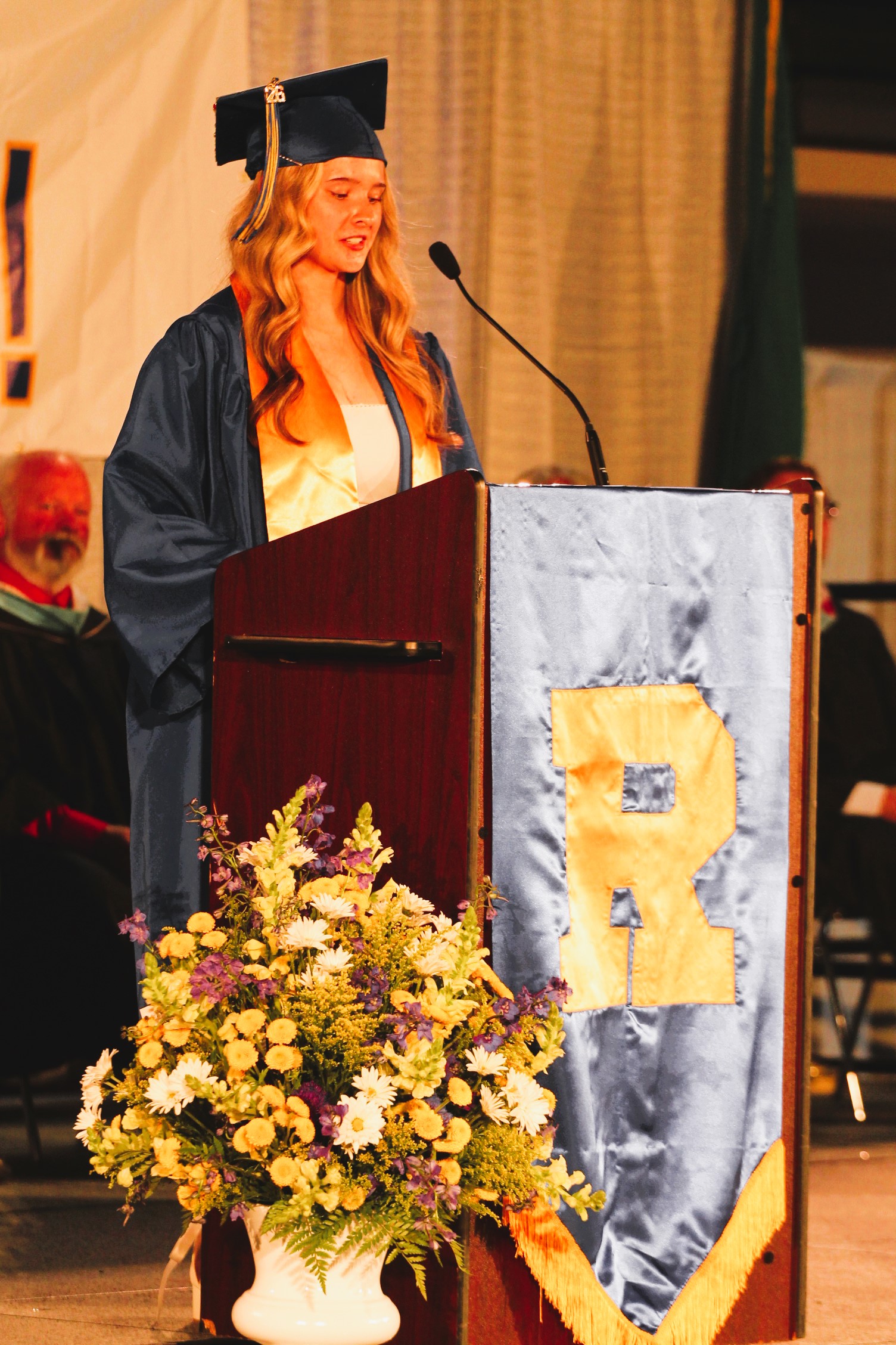 A woman in a cap and gown stands at a podium with a microphone, a banner, and flowers.