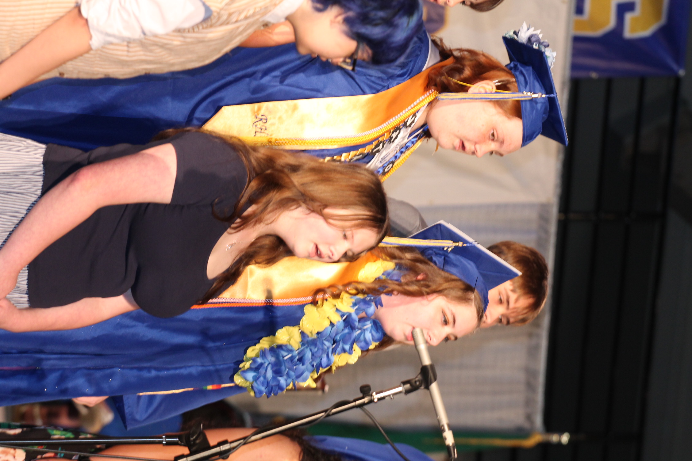 Several people in caps and gowns stand on a stage, two of them singing into a microphone.