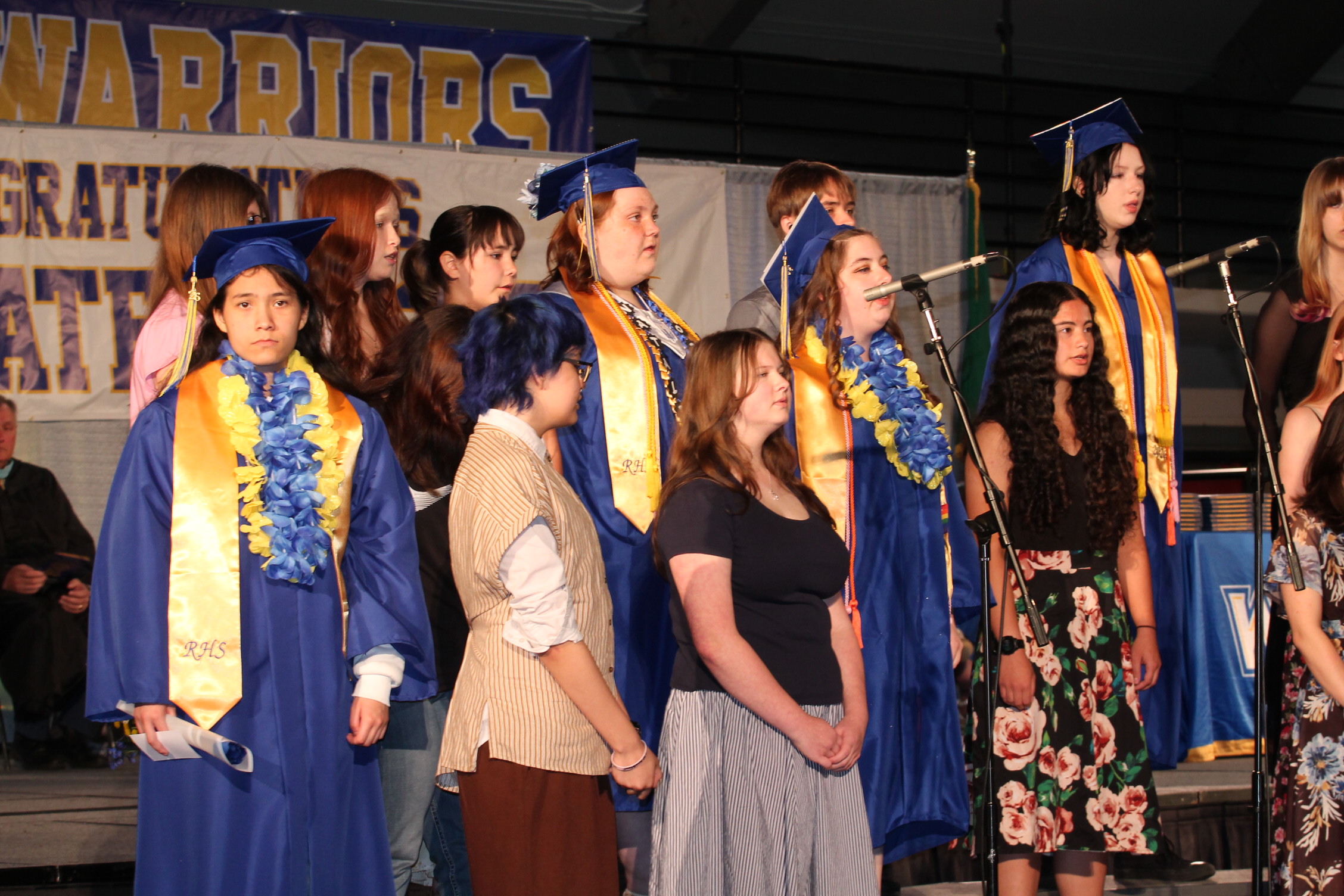 A group of students wearing blue gowns and caps, some with yellow sashes, standing on a stage.
