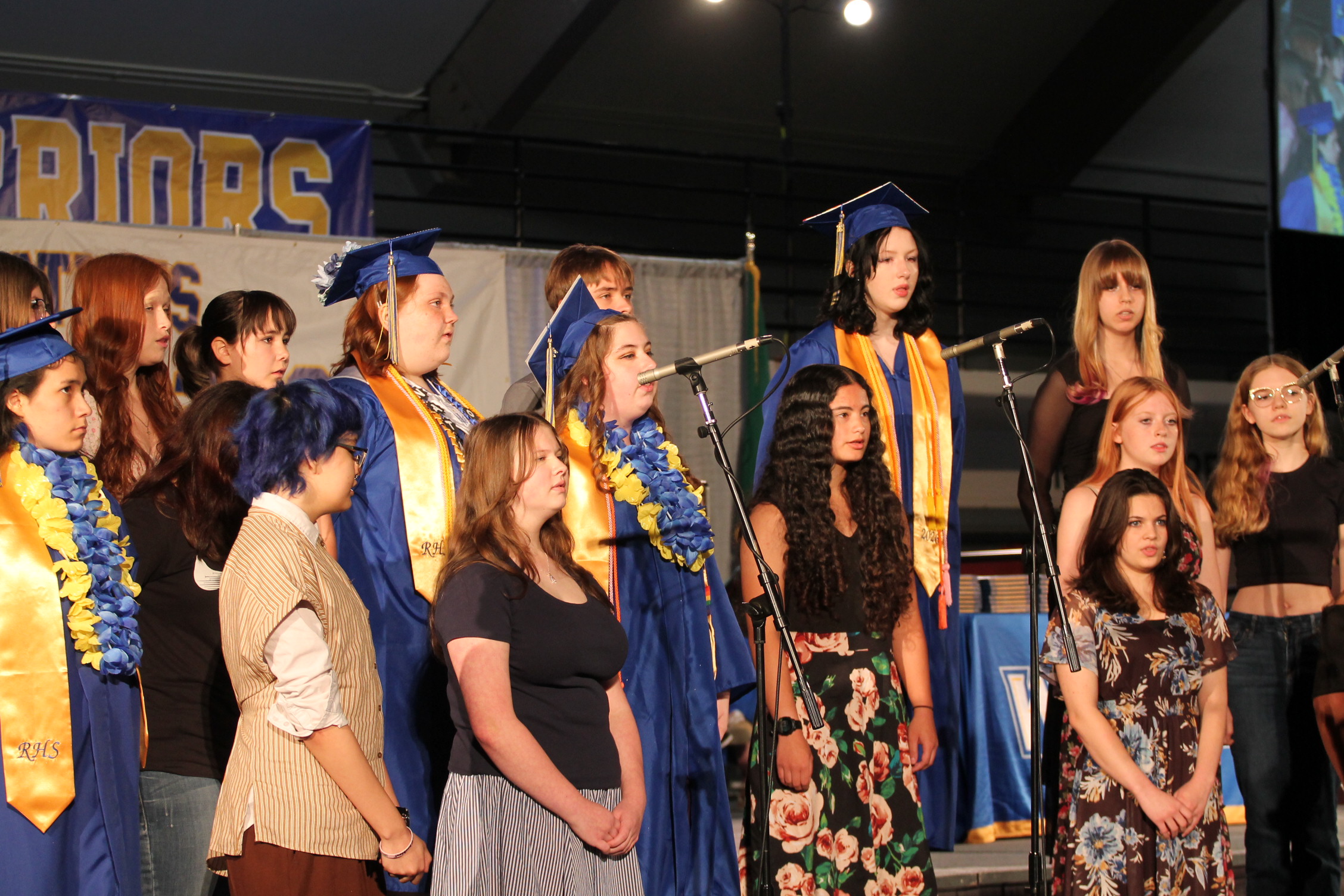 A choir stands on a stage, all dressed in blue gowns and some with graduation caps. They face a banner and microphones.