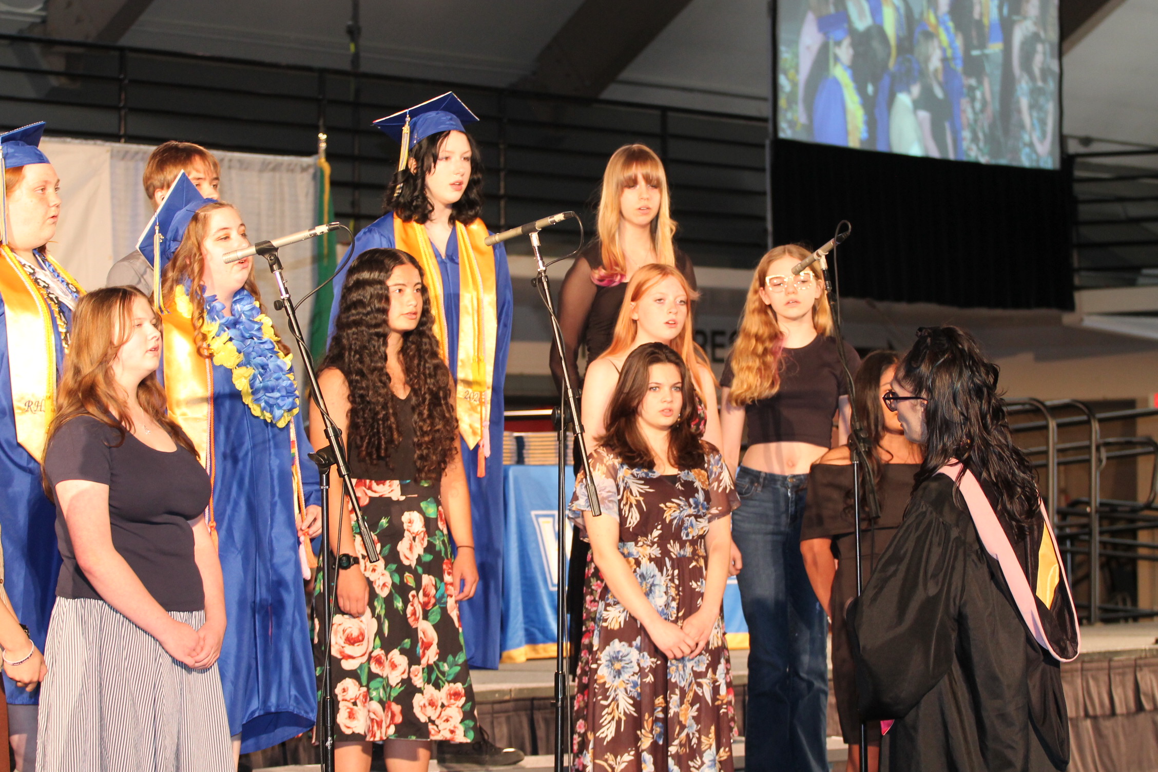 A group of young women in gowns and caps, standing on stage, with microphones and a large screen in the background.