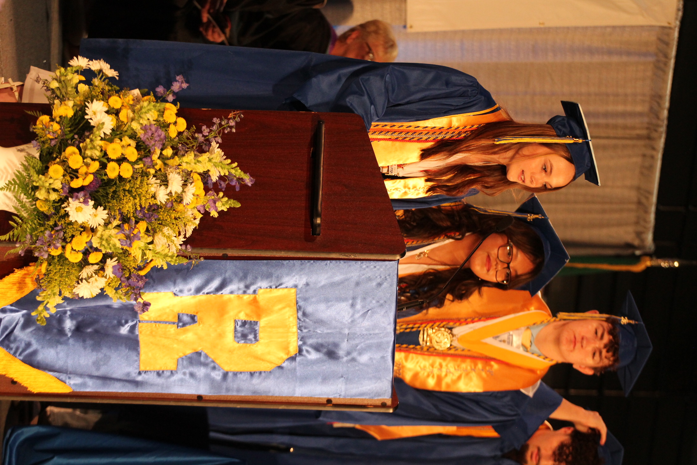Four individuals, in caps and gowns, stand at a podium during a ceremony. A flower arrangement is placed in front.
