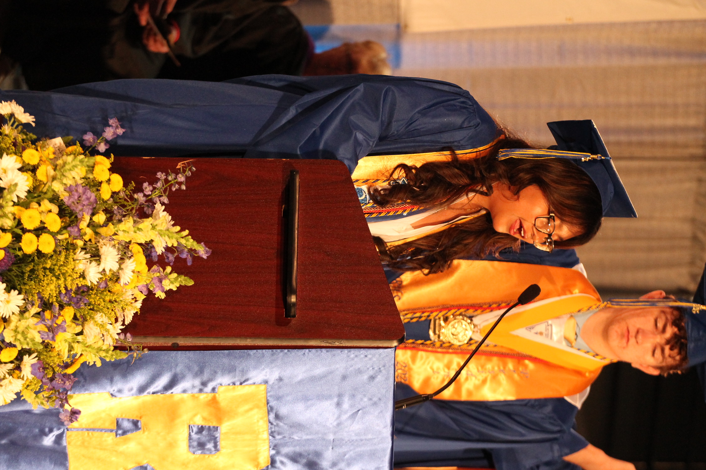 A woman in a blue gown speaks at a podium with a microphone. A man stands behind her, wearing a yellow gown. An audience sits in the background.