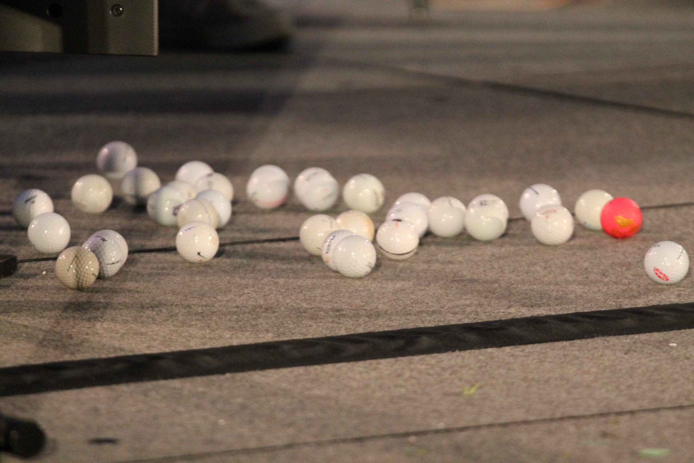 Multiple white golf balls with one red one scattered on a concrete floor with a black line.