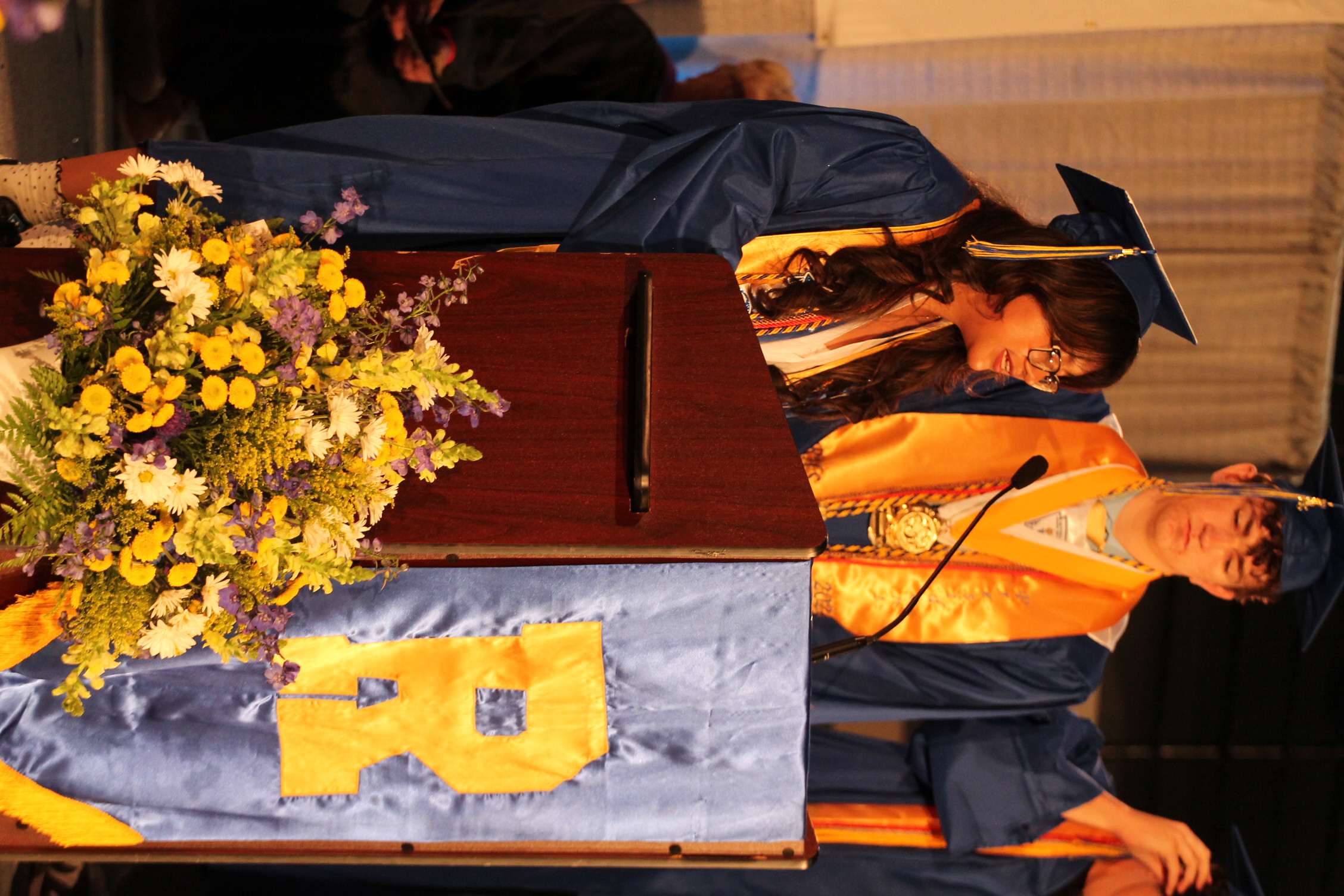 Woman speaking at podium with flowers at front; a man behind her wearing a graduation gown.