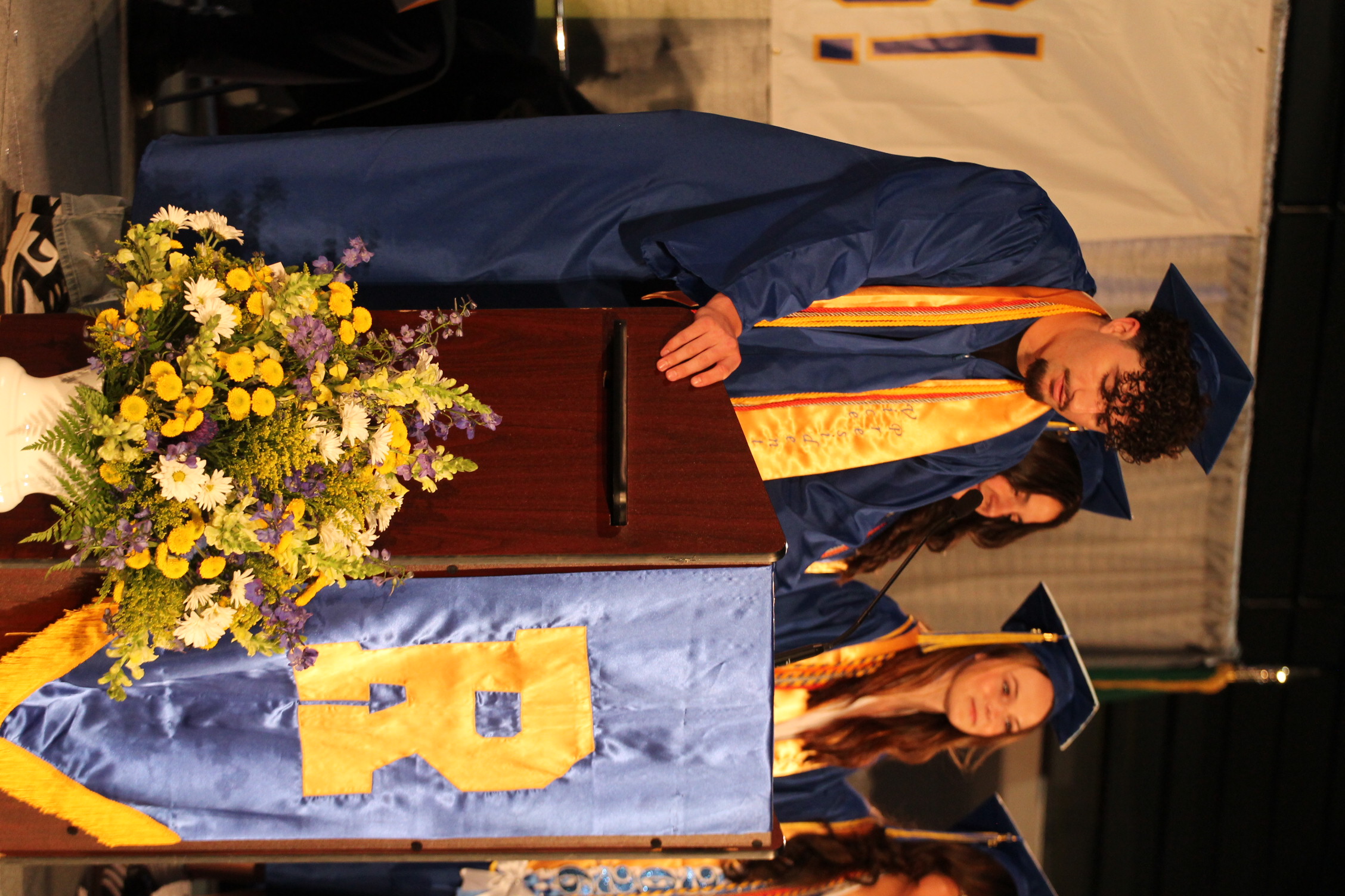 A man and woman in graduation robes stand at a podium. The man holds the podium, and the woman stands behind it.