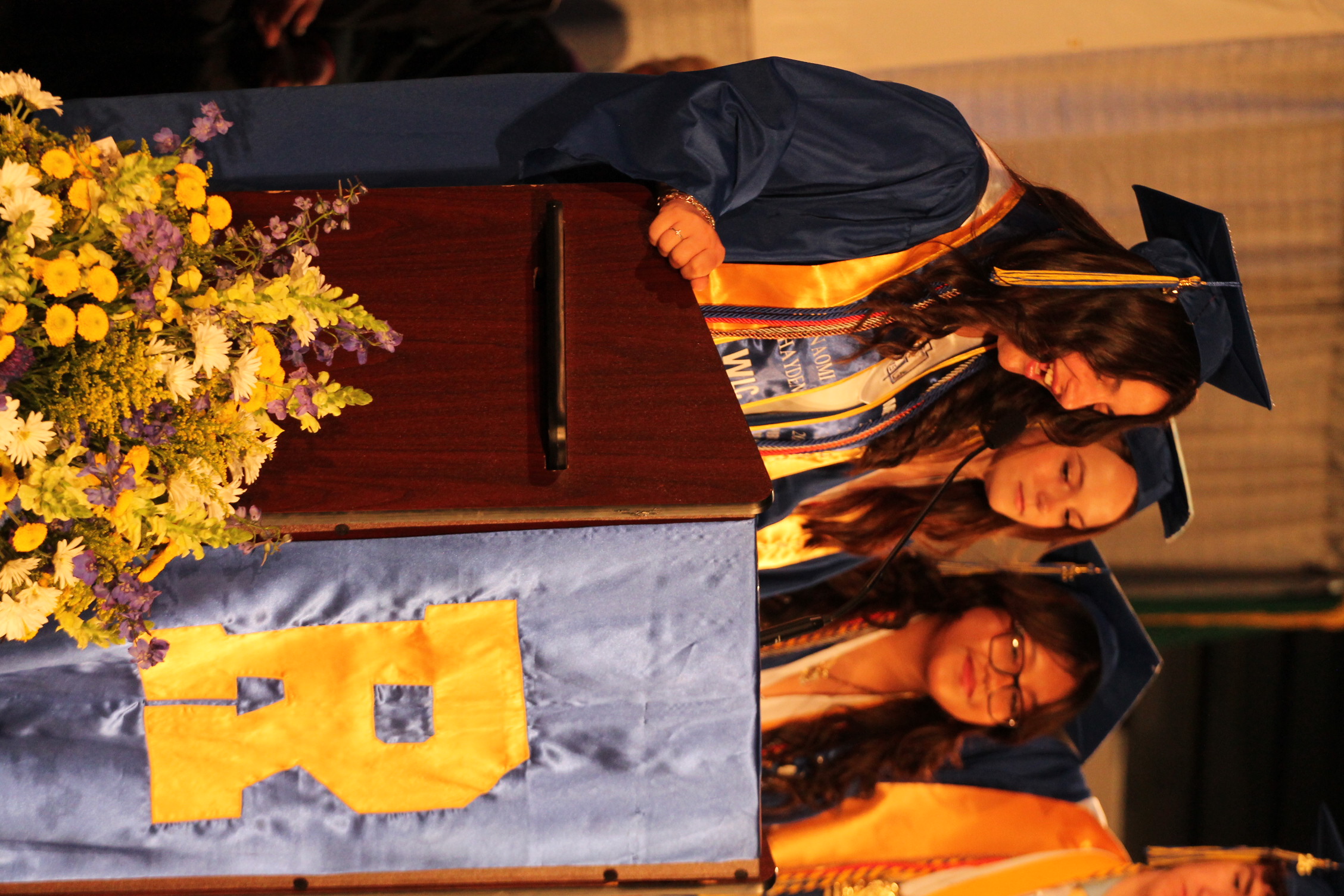 Three students in graduation gowns stand at a podium with a blue banner reading "R" and a bouquet of flowers.