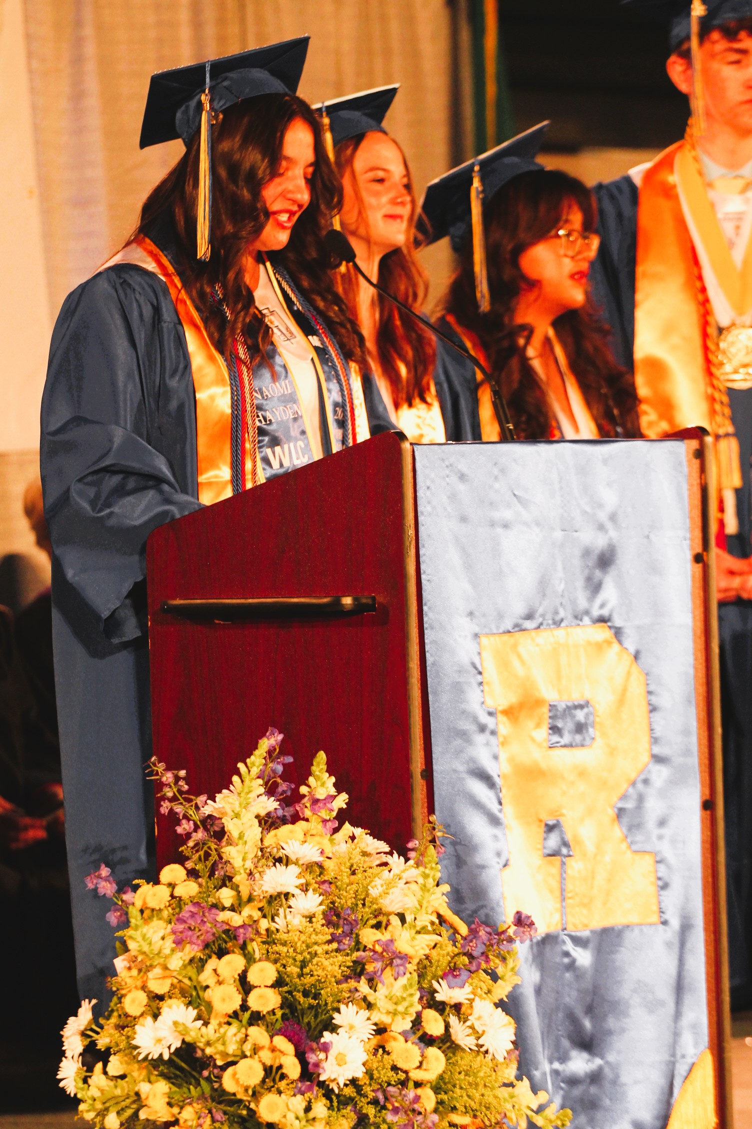 Graduates wearing caps and gowns, two women speaking into microphones, a podium with flowers and a banner reading "R".